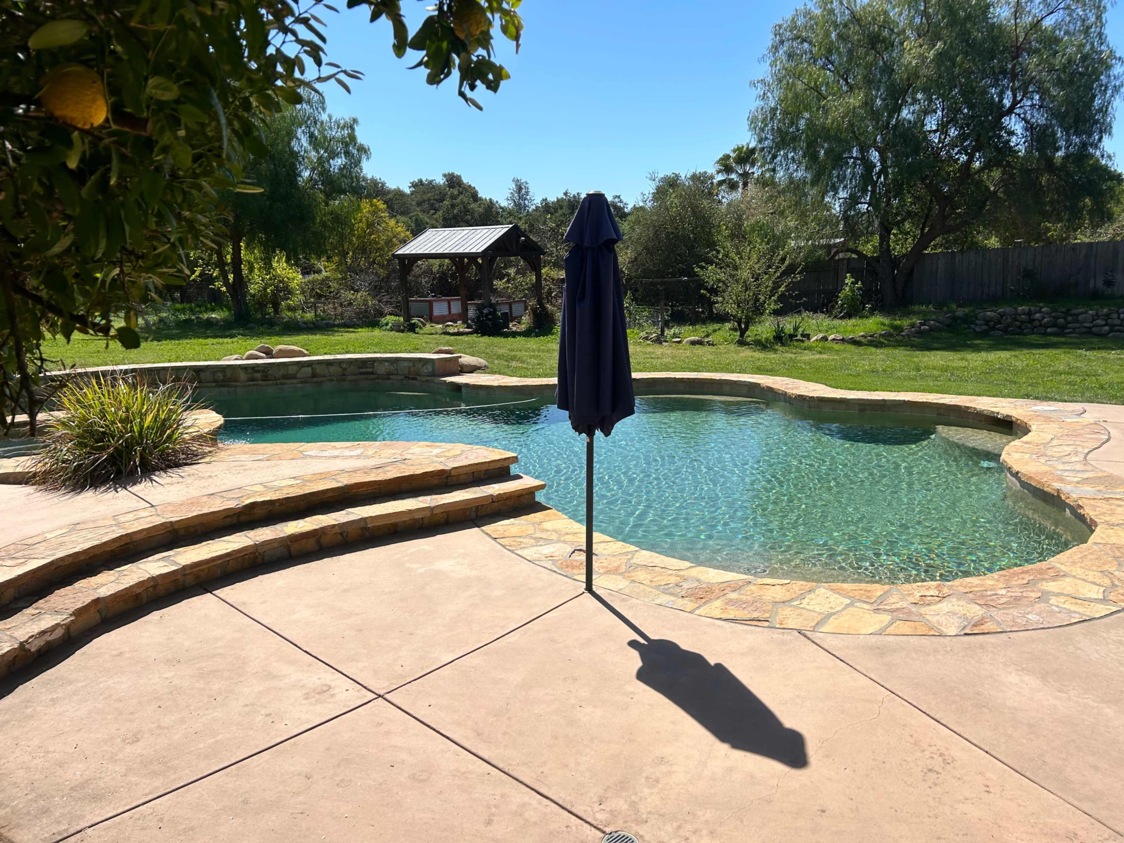 A swimming pool with a stone patio and a blue umbrella is surrounded by lush greenery and a gazebo in the background.