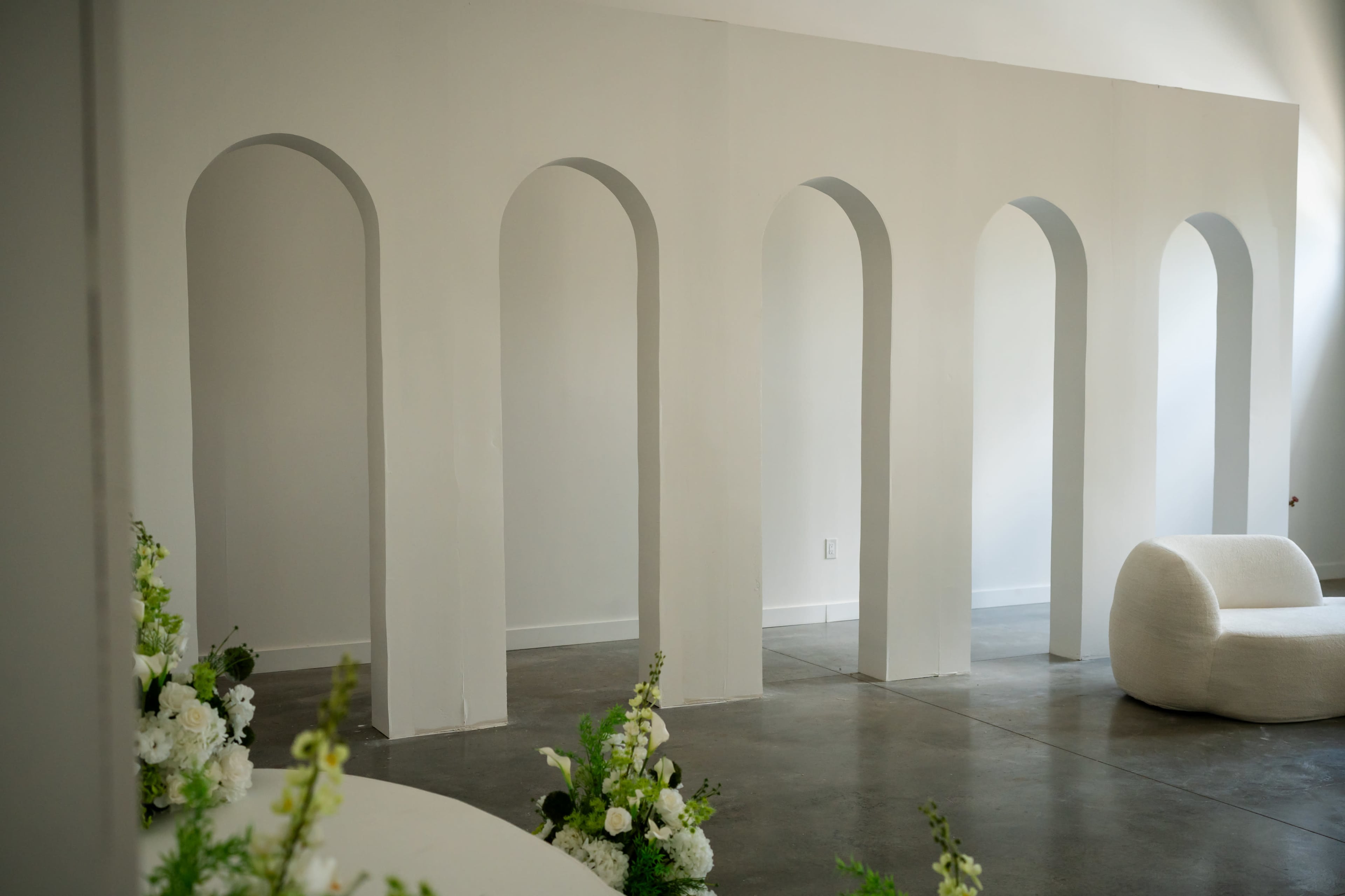The image shows a minimalistic interior space featuring a series of white archways and a low, curved seating area, with floral arrangements at the base of the archways.