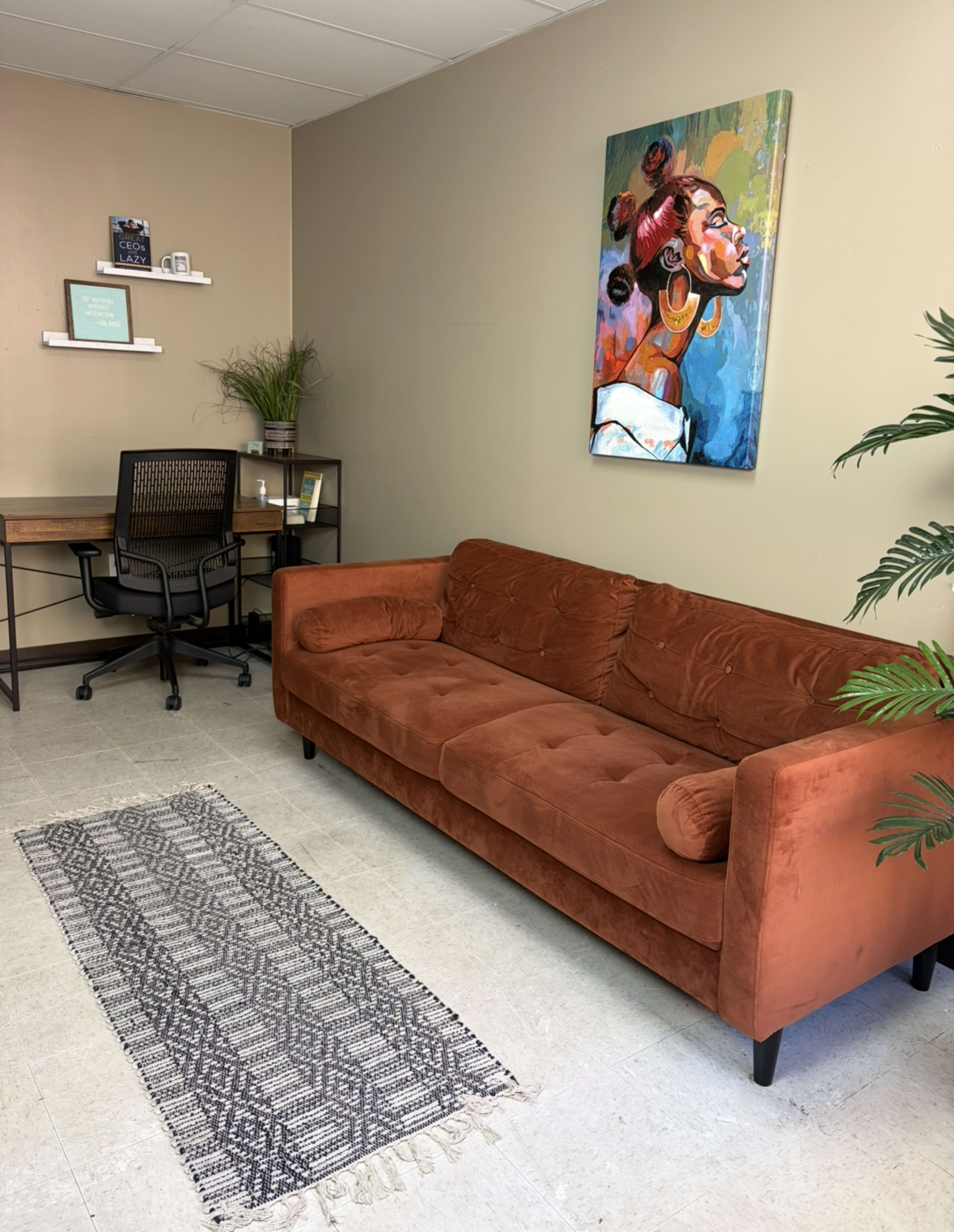 The image shows a small office space featuring a brown couch, a patterned rug, a desk with a chair, and a colorful painting on the wall.