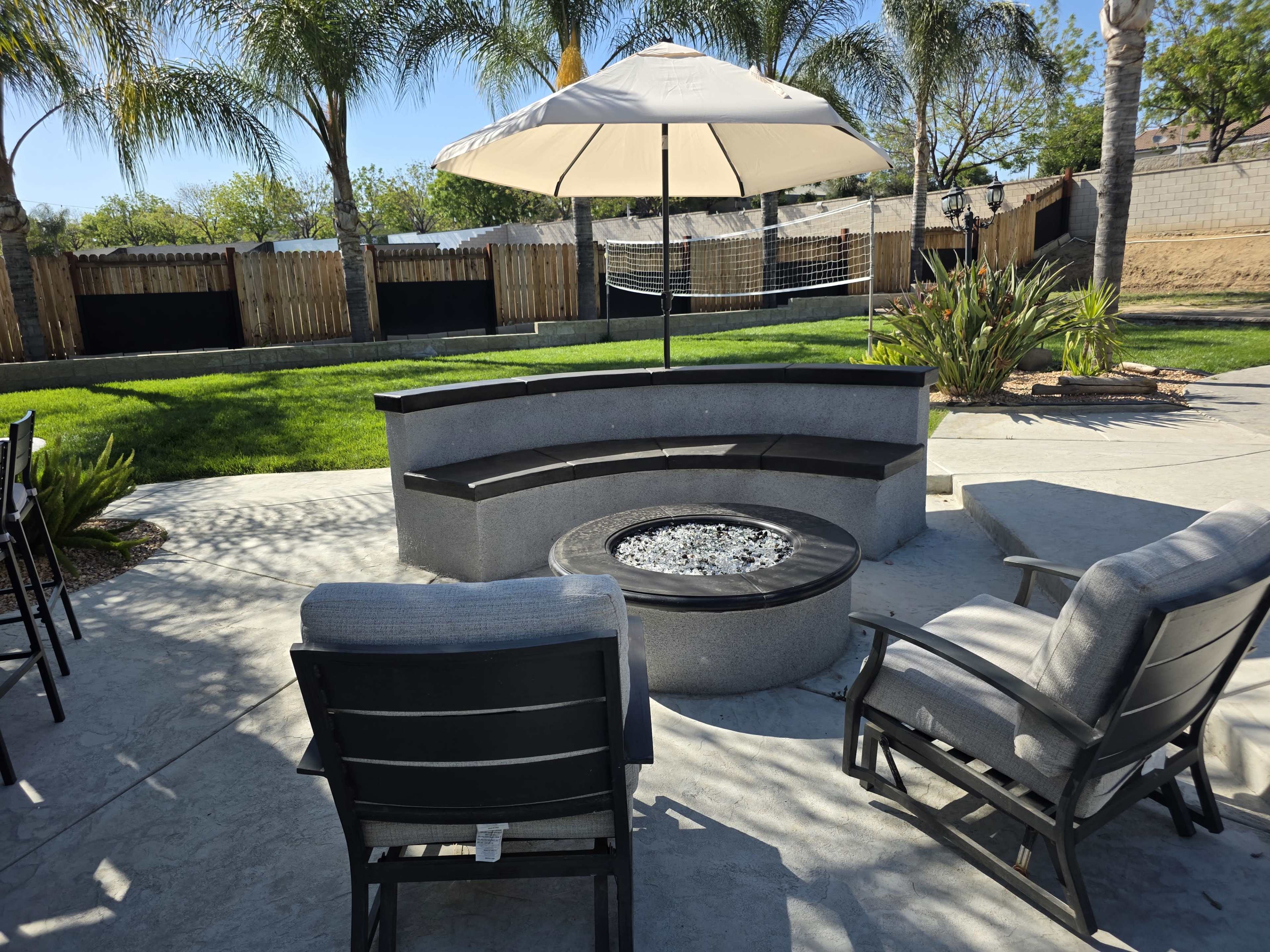 The image shows a circular stone fire pit surrounded by several outdoor chairs and an umbrella, set against a backdrop of grass and palm trees.