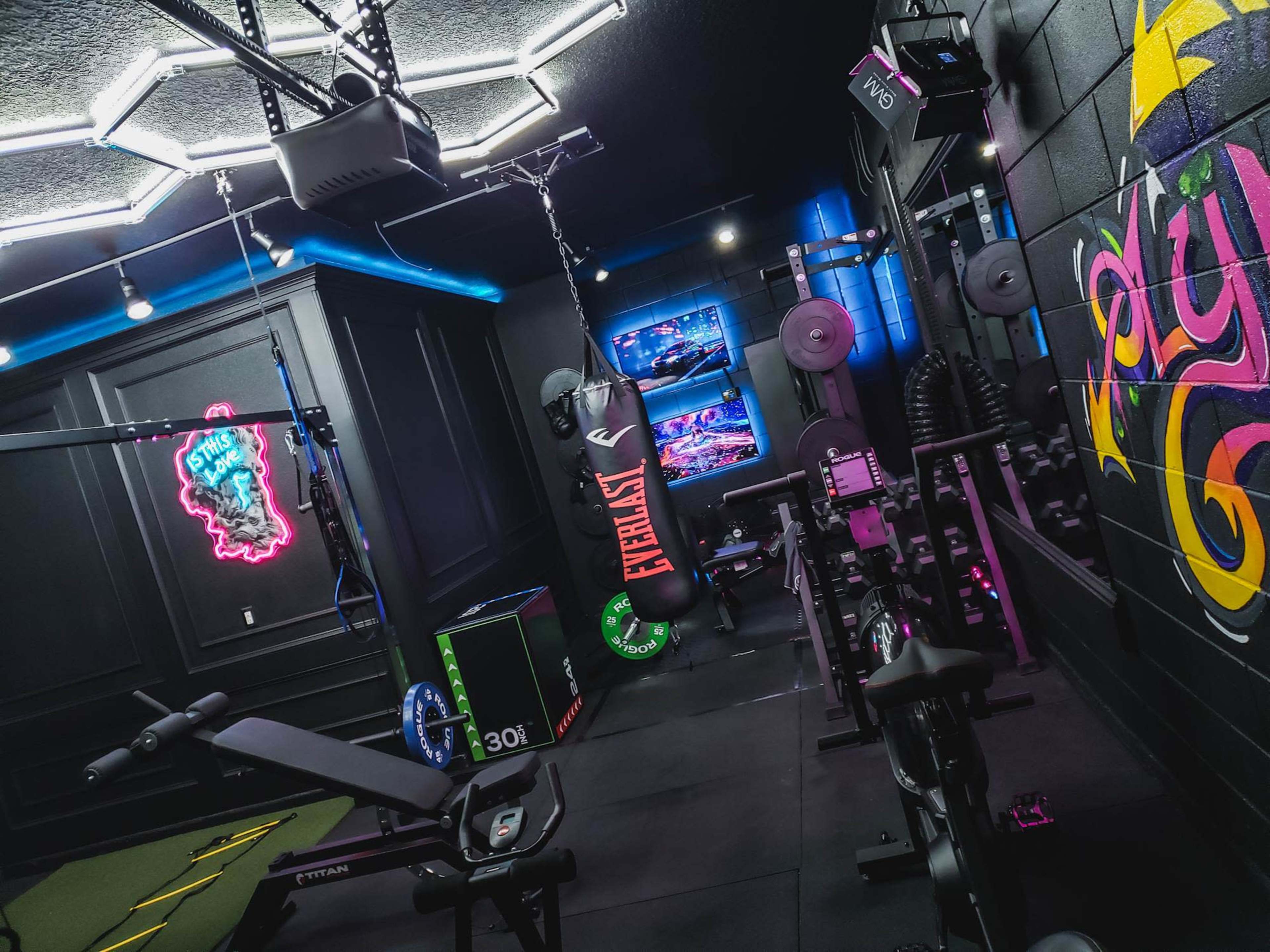 The image shows a well-equipped home gym featuring exercise machines, weightlifting equipment, a punching bag, and colorful neon lighting.