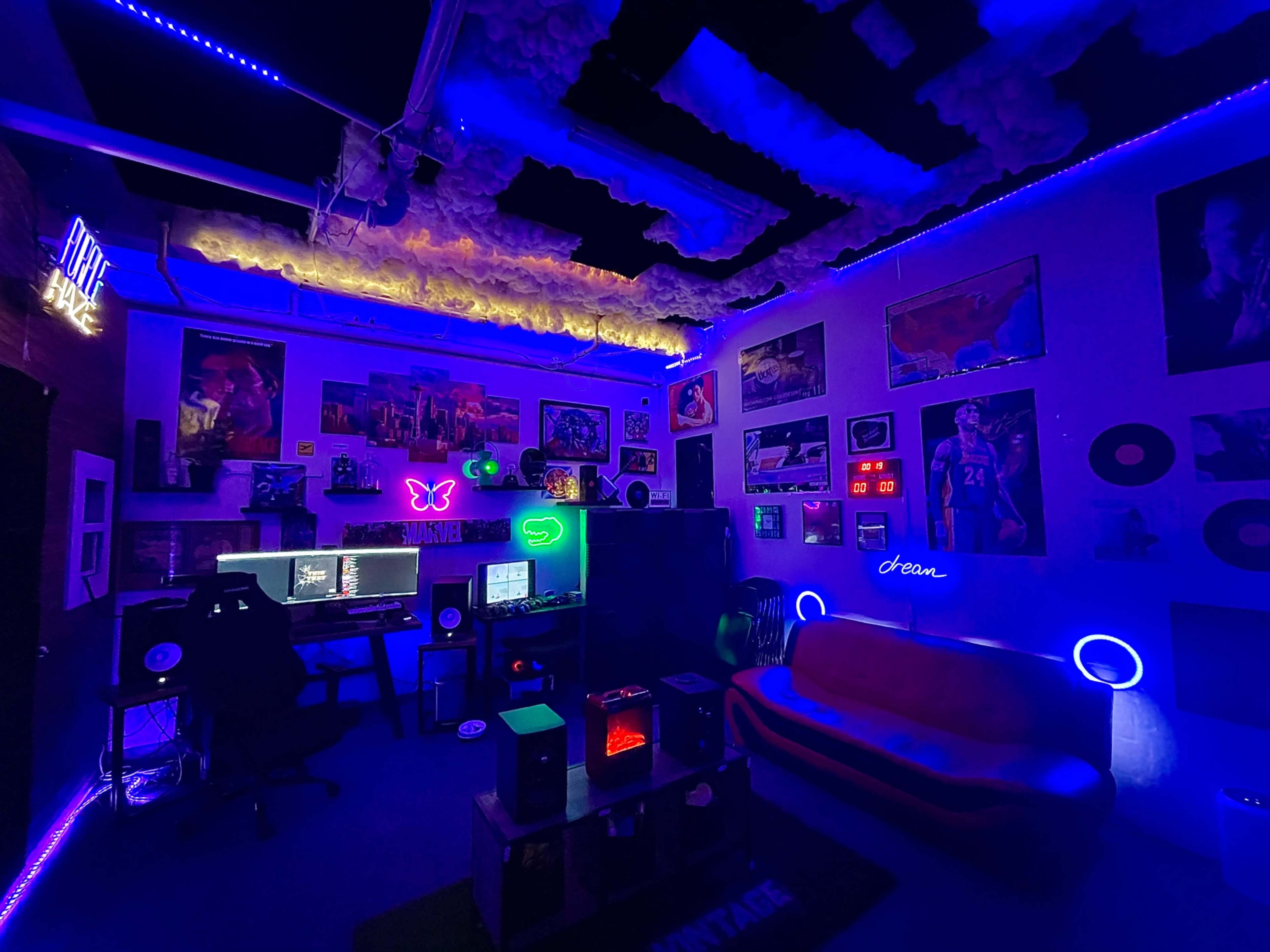 The image shows a dimly lit room decorated with colorful neon lights, a computer setup with multiple monitors, vinyl records on the walls, and a couch.