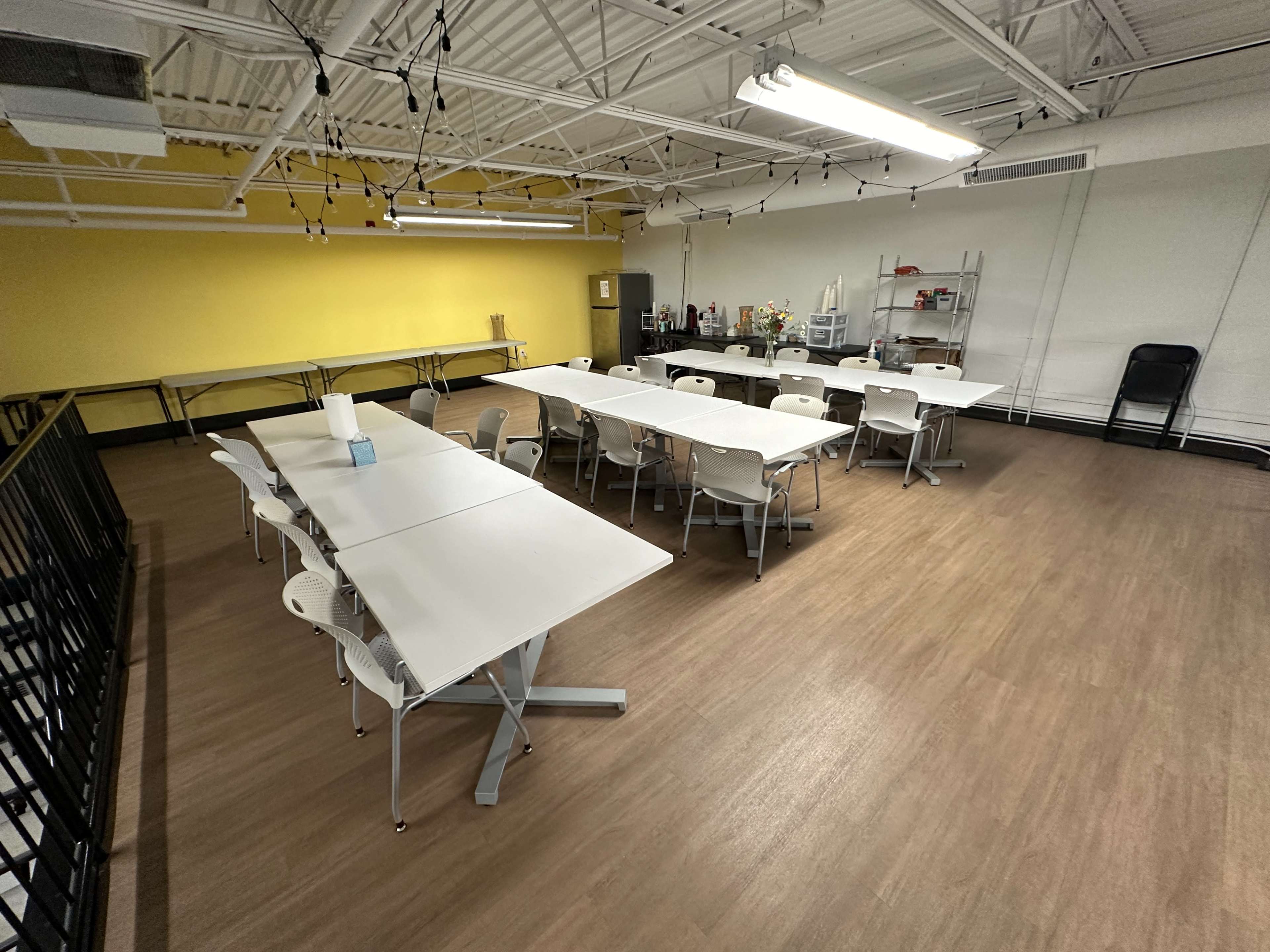 A spacious, well-lit classroom features several white tables and chairs arranged around the room with a yellow wall in the background.