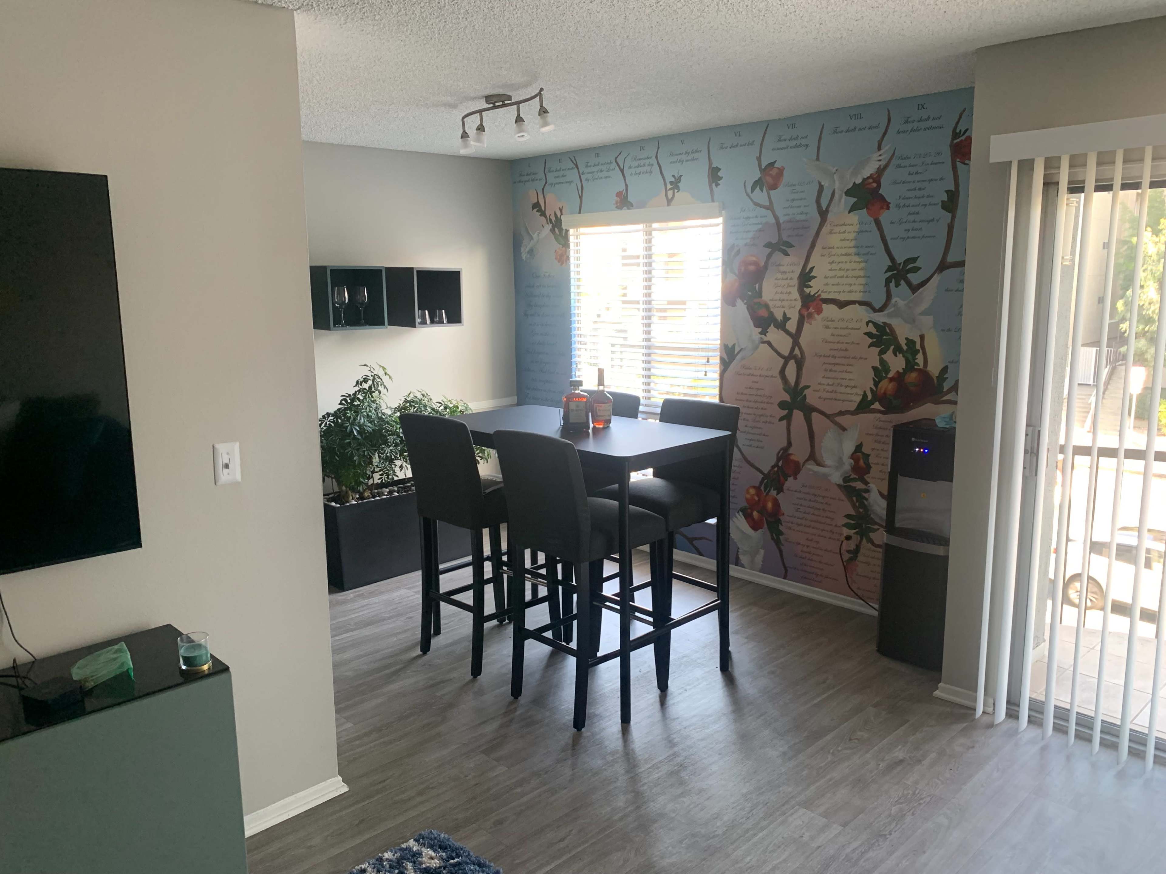A living space featuring a dining area with tall black chairs around a square table, a wall adorned with a colorful mural, and a large window allowing natural light to enter.
