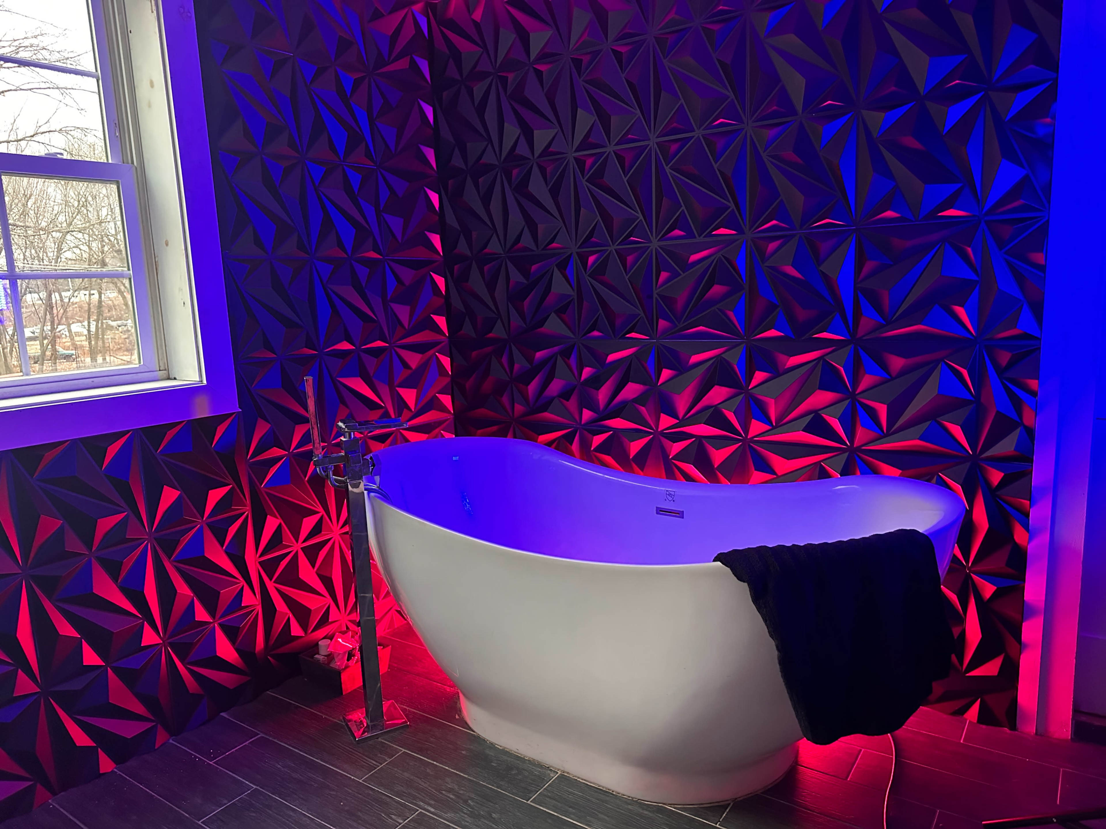A modern bathroom featuring a sleek white freestanding bathtub beside a window, with a textured wall illuminated in red and blue lights.
