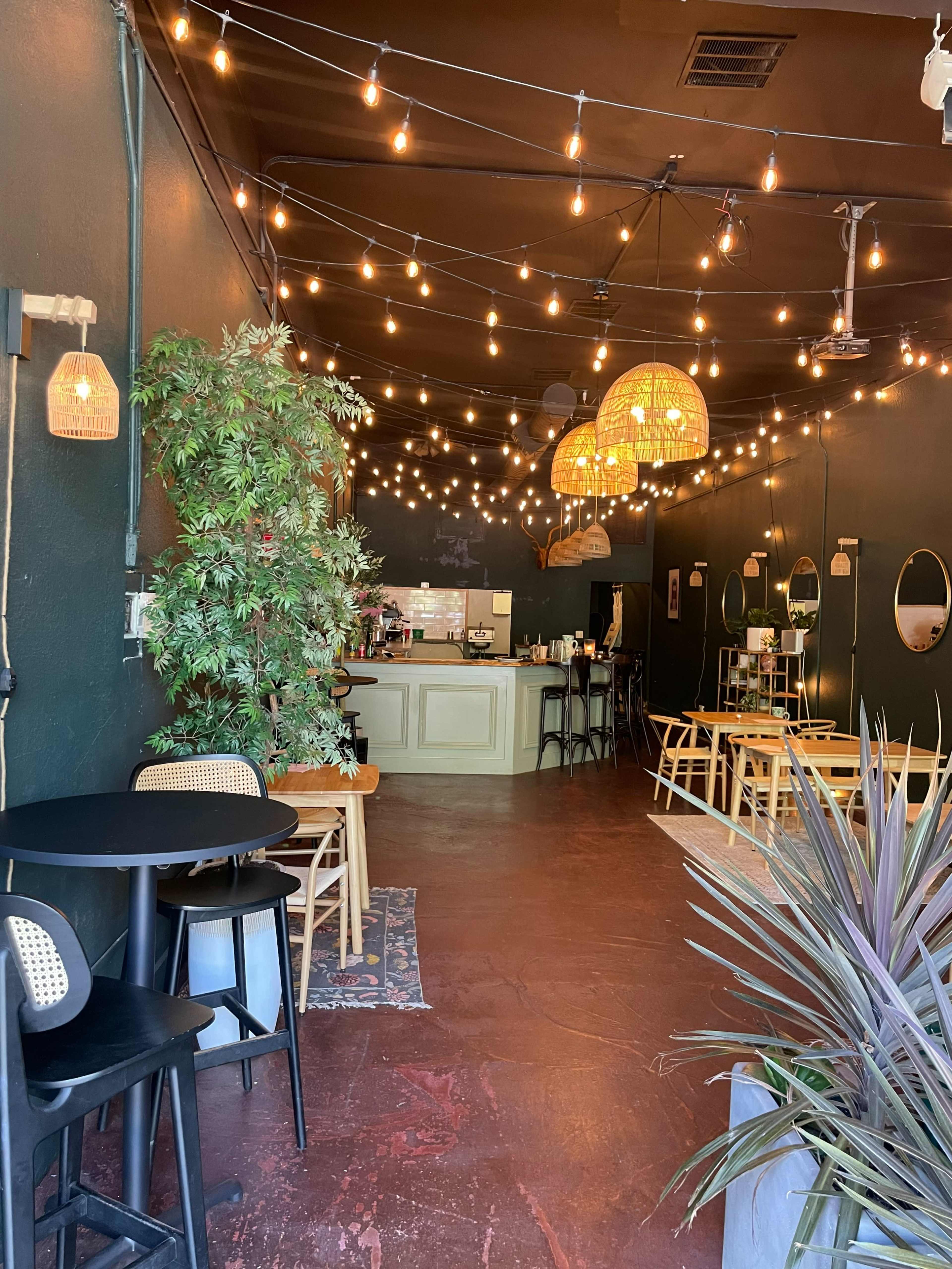 A cozy café features a dark green interior, string lights overhead, and a mix of seating arrangements, including tables and chairs.