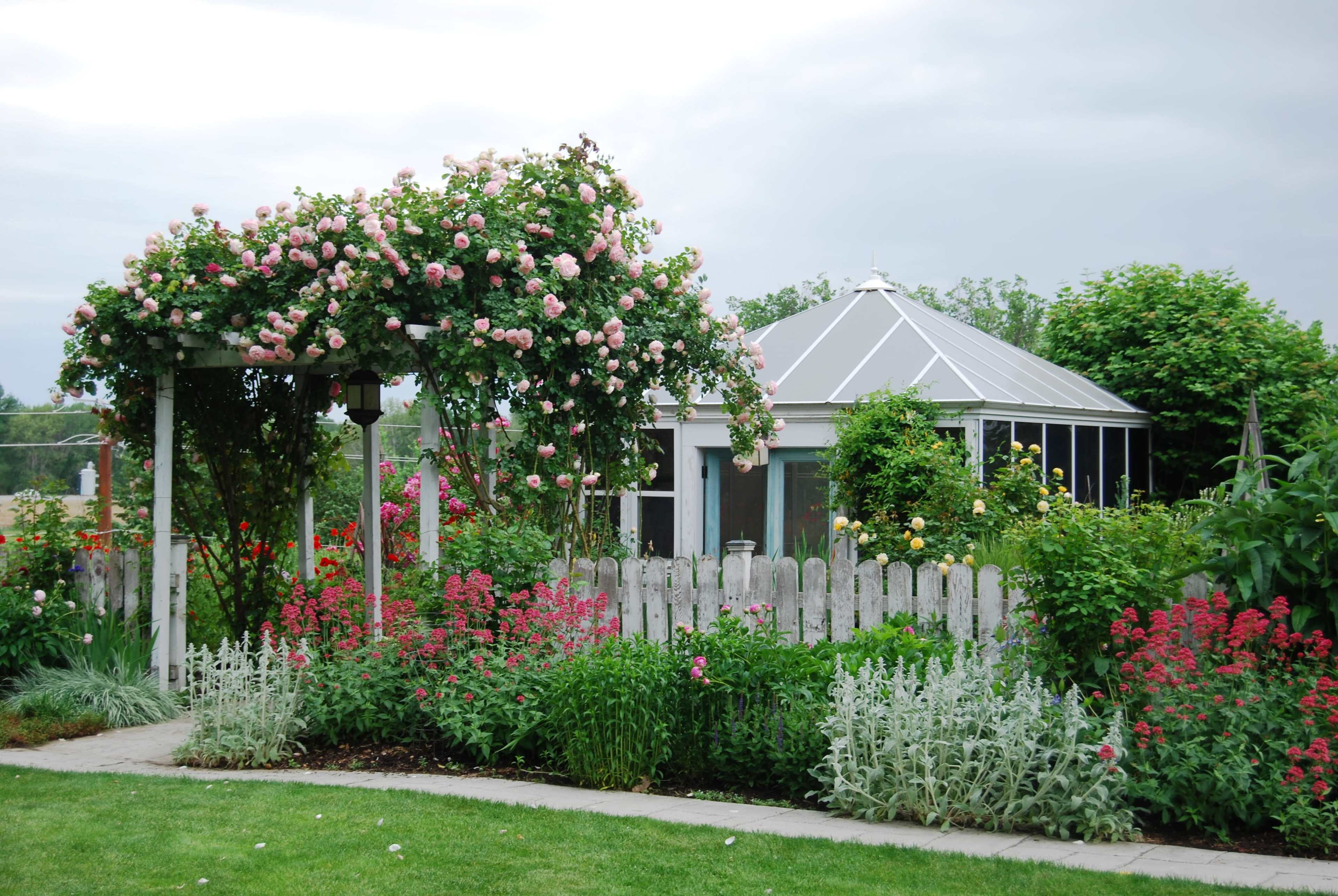 A gazebo adorned with blooming roses stands near a house surrounded by a colorful garden and a white picket fence.