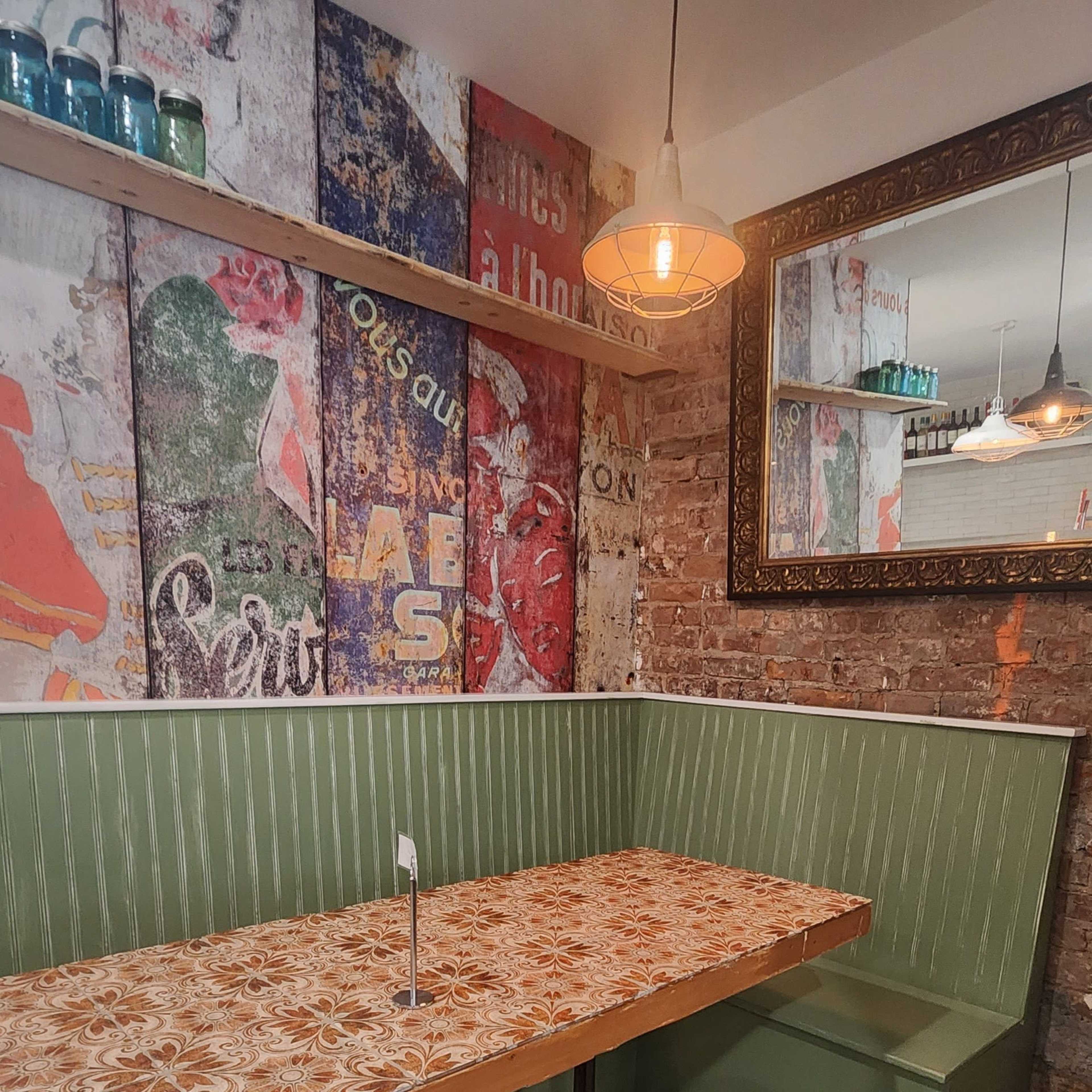 The image shows a cozy restaurant corner featuring a patterned table, green seating, and a vintage mural on the wall.