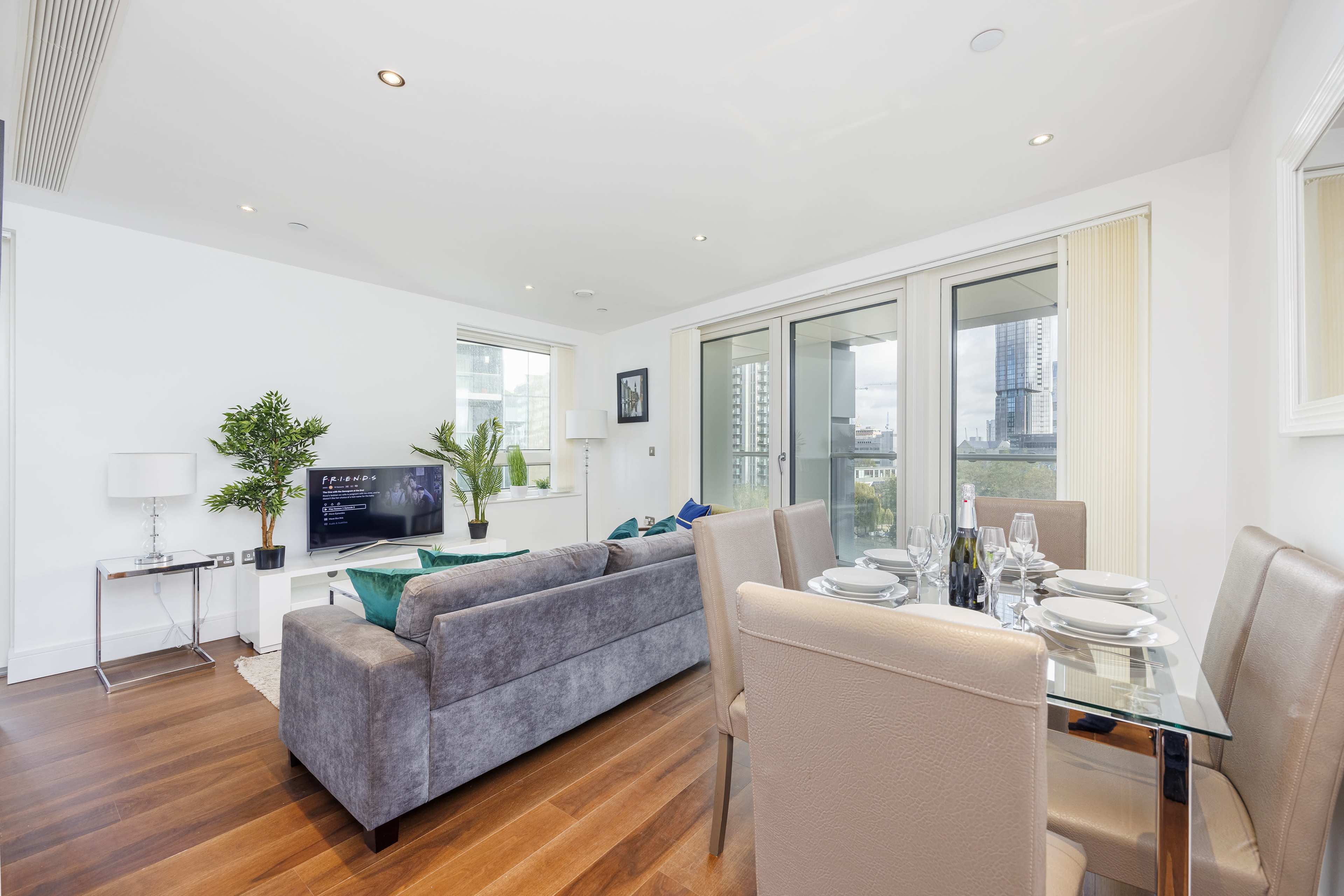 A modern living area with a gray sofa, a dining table set for four, and large windows providing a view of a city skyline.