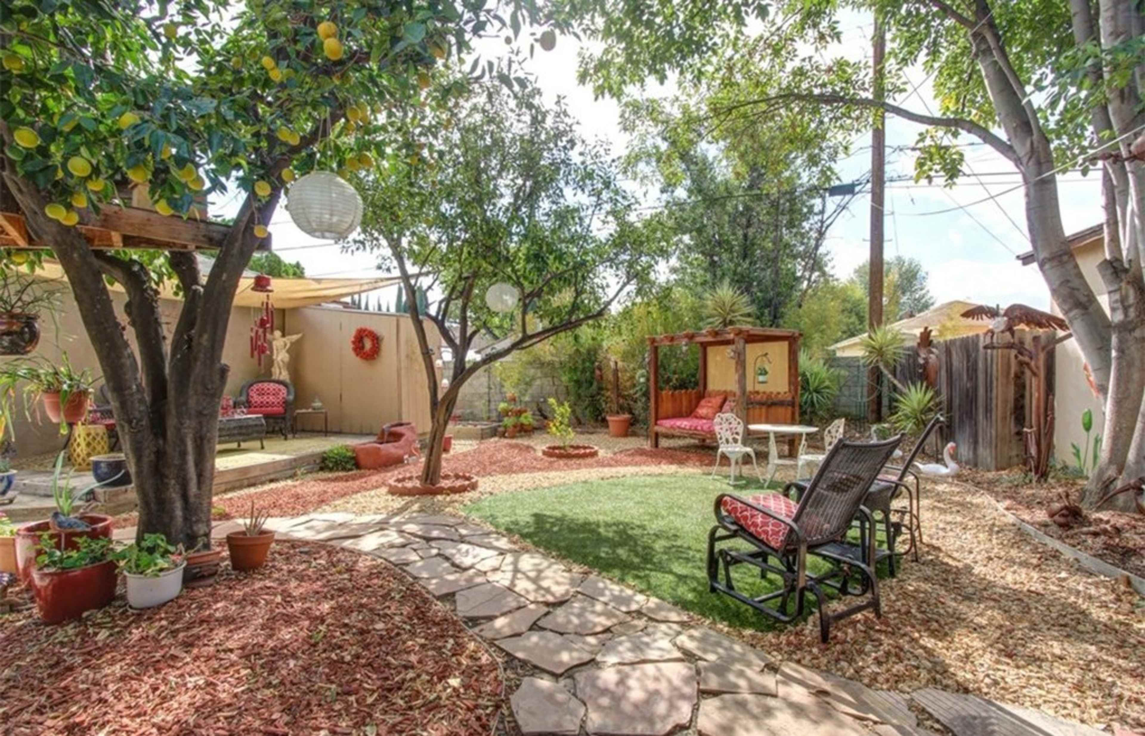 The image shows a landscaped backyard featuring stone pathways, a green lawn, potted plants, and a seating area with chairs and a swing under trees.