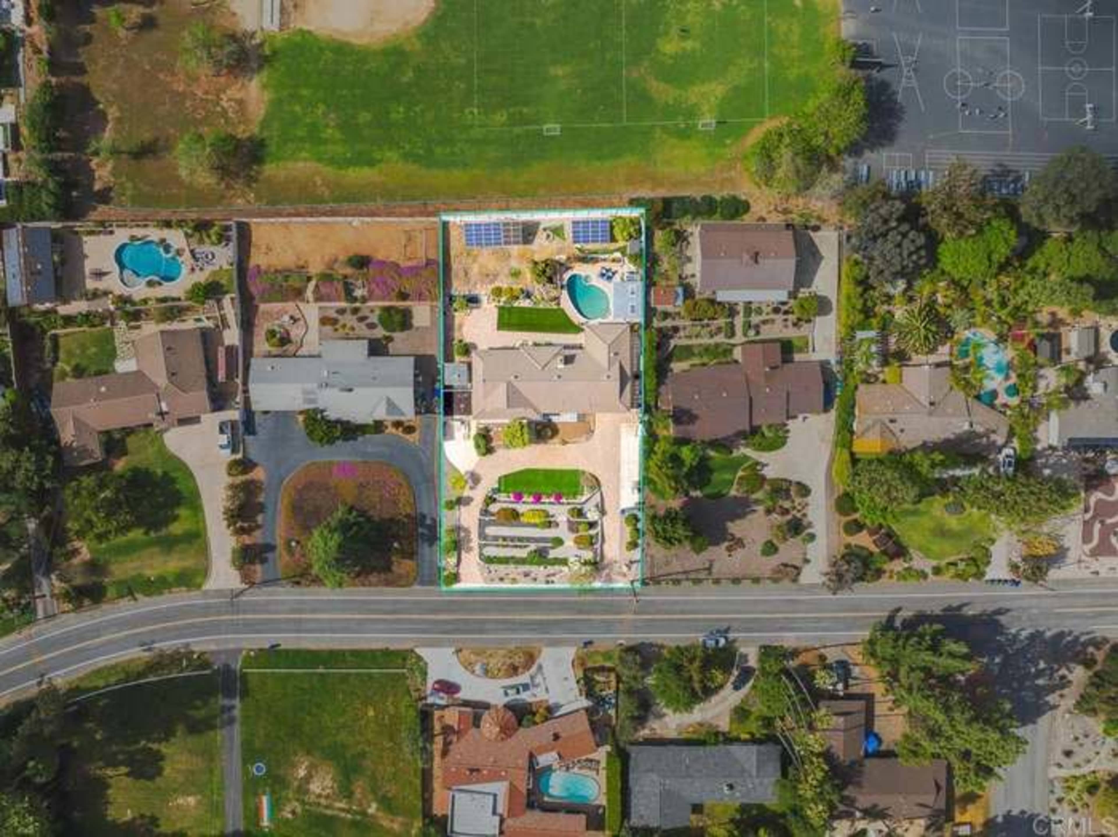 The aerial view shows a residential area with several houses, a large yard featuring a pool and landscaped gardens, and a nearby sports court.