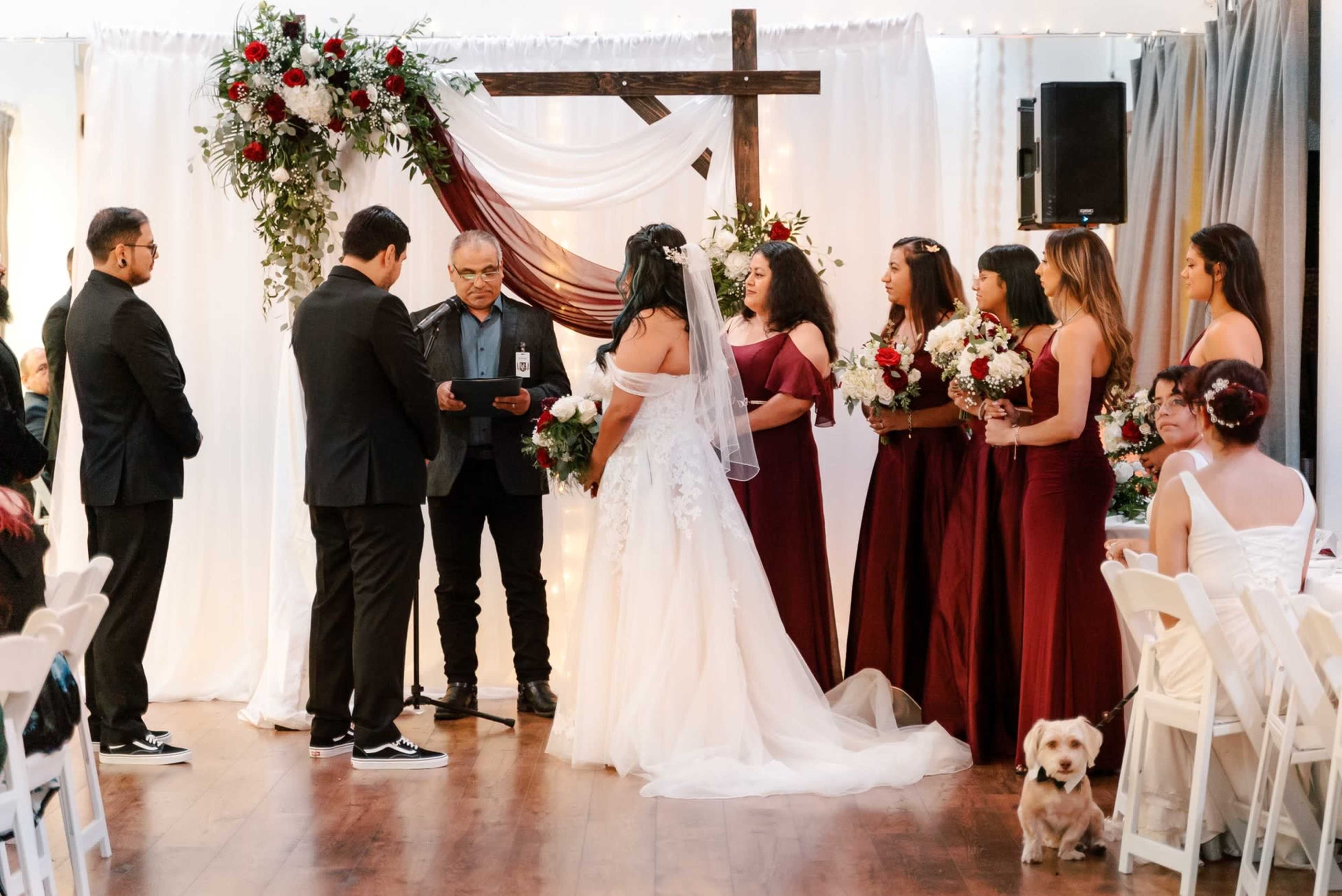 A wedding ceremony is taking place indoors with a decorated altar featuring flowers and a cross, as the bride and groom stand before an officiant surrounded by a wedding party.