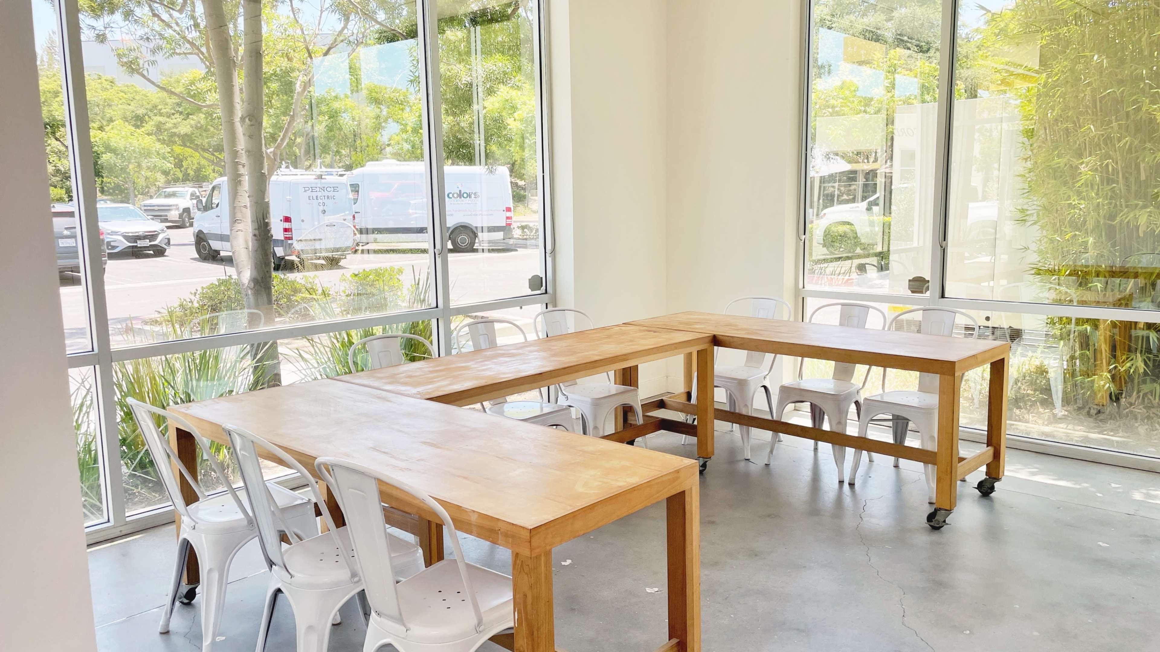 A bright room features two wooden tables arranged in an L-shape, surrounded by several white chairs, with large windows revealing greenery and parked vehicles outside.
