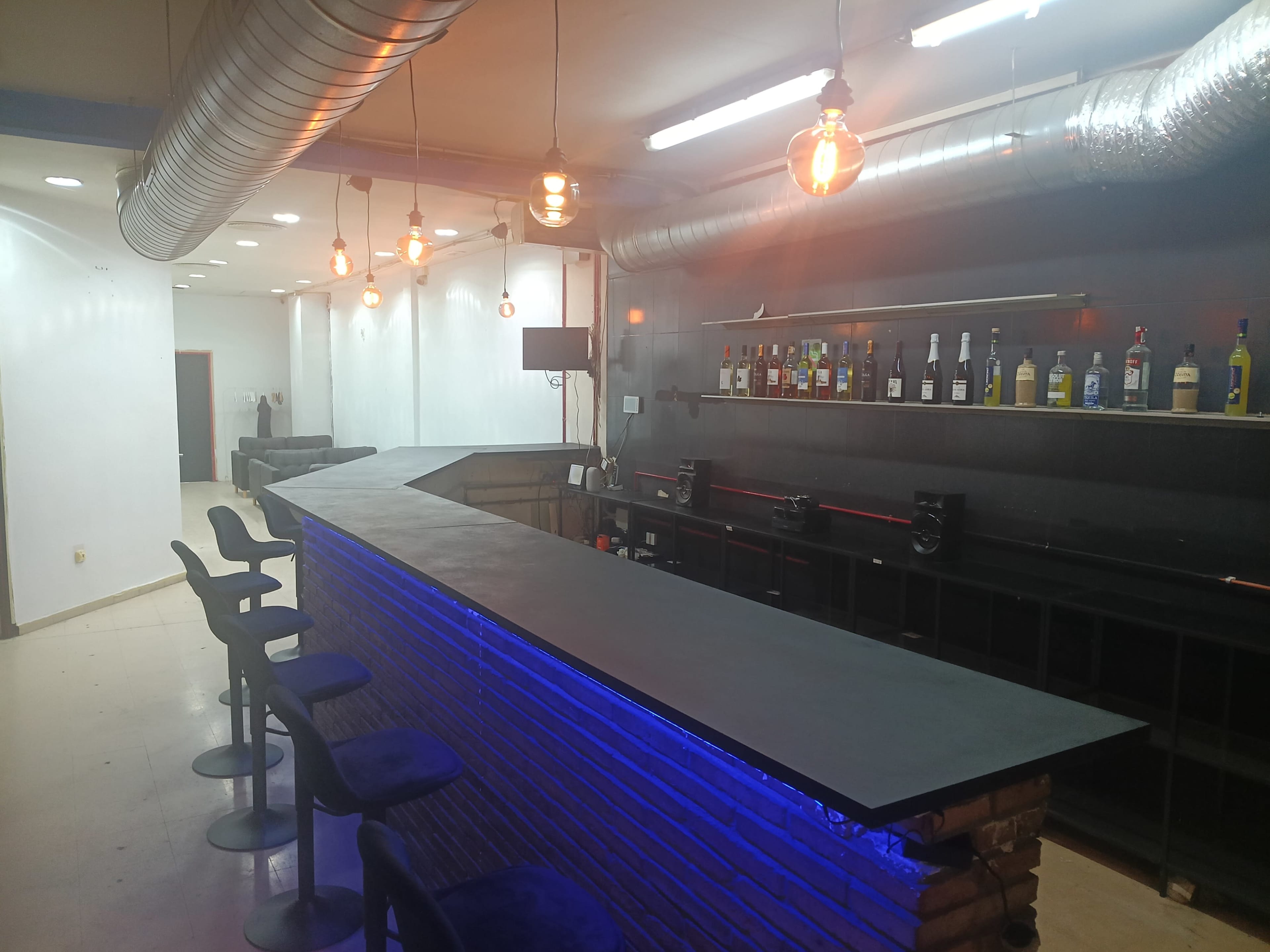 The image shows a modern bar interior featuring a curved countertop with blue LED lighting, surrounded by high stools and a backdrop of various liquor bottles on shelves.