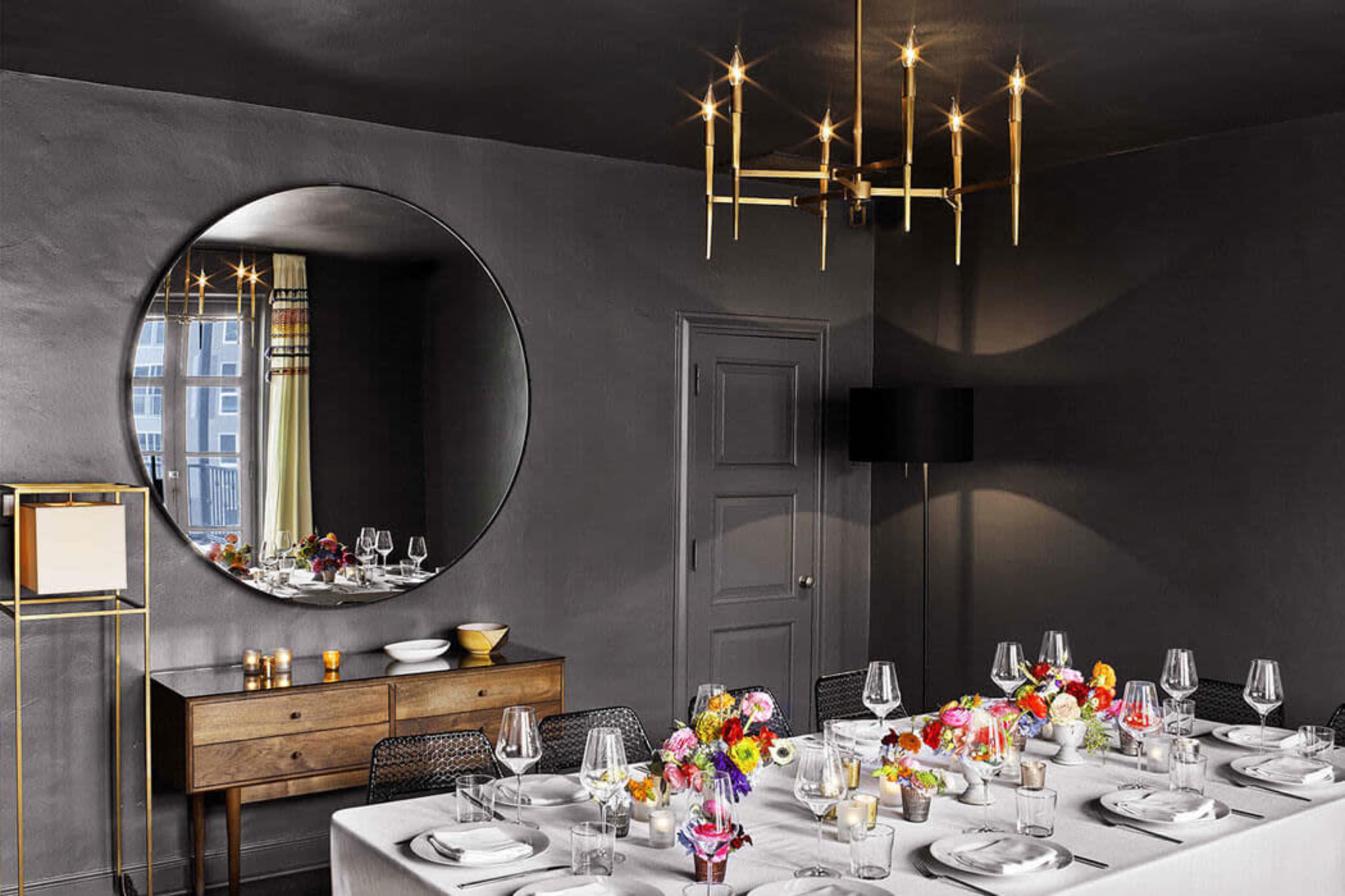 A modern dining room features a large round mirror, a minimalist chandelier, and a table set with elegant tableware and a colorful floral centerpiece.