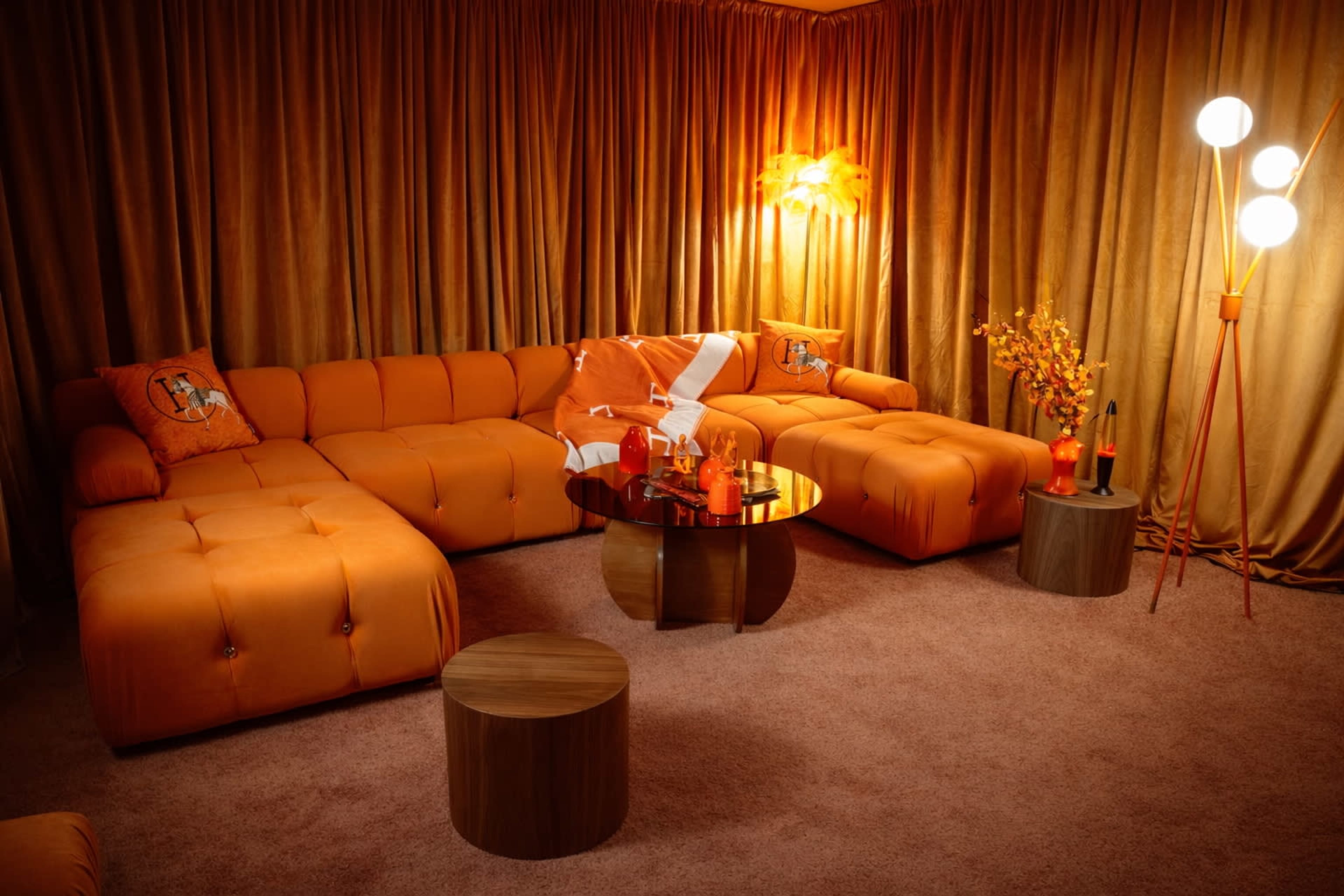 The image features a cozy living room with an orange sectional sofa, a round glass coffee table, and decorative elements like a lamp and a flower arrangement, all set against gold-toned curtains.
