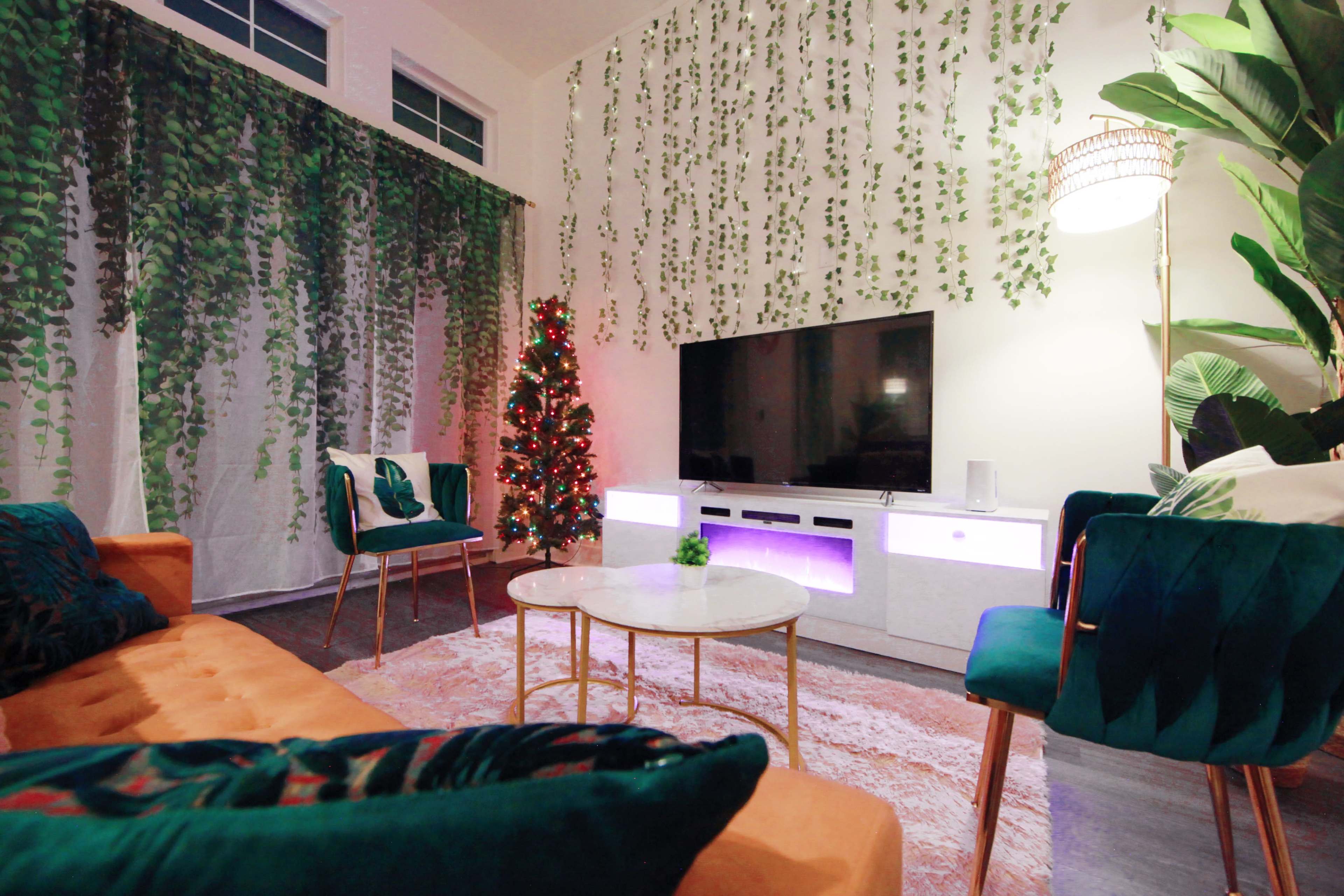 The living room features a television mounted on a white cabinet, surrounded by green plants and a small decorated Christmas tree, with modern furniture arranged around a low coffee table.