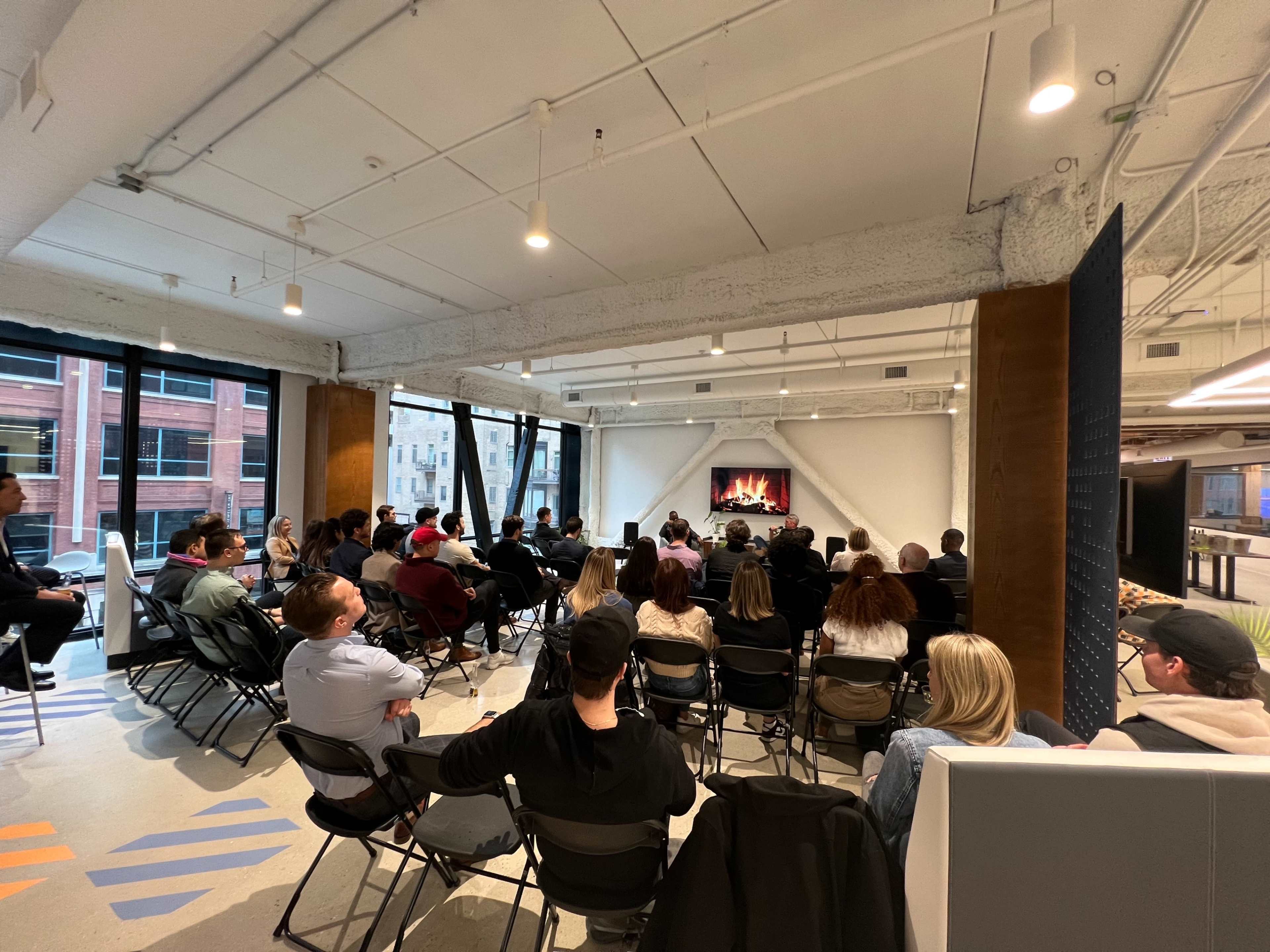 A group of people is seated in an indoor space for a presentation, facing a screen that displays a fireplace.