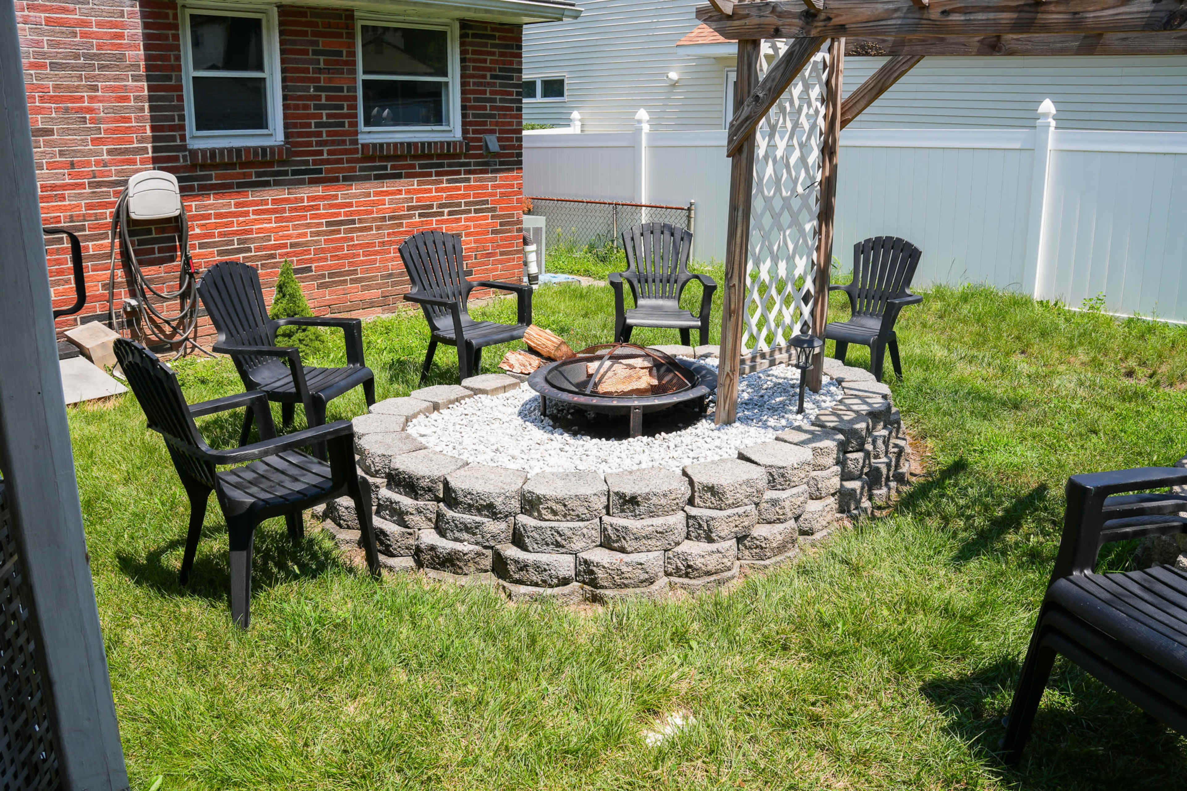 A circular stone fire pit surrounded by several black chairs is situated on a grassy area in a backyard.