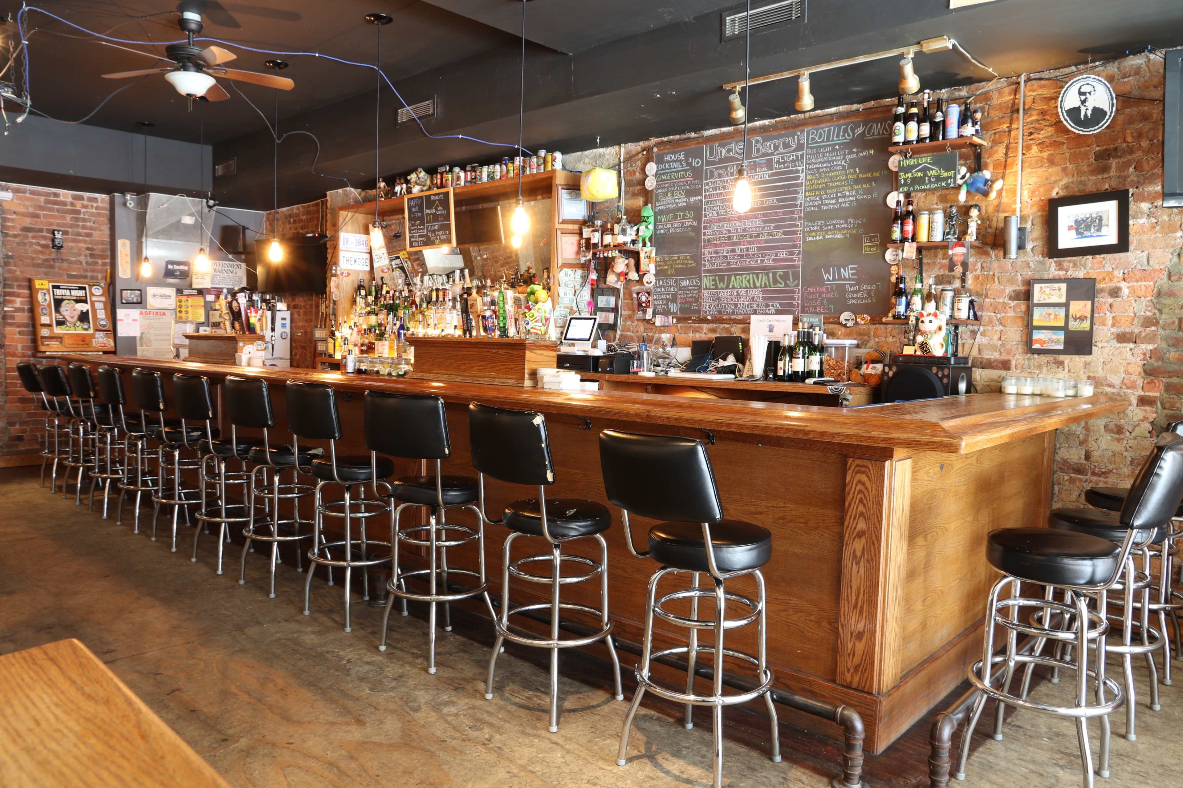 A spacious bar with a wooden counter lined with black stools, surrounded by exposed brick walls and shelves filled with various bottles of alcohol.