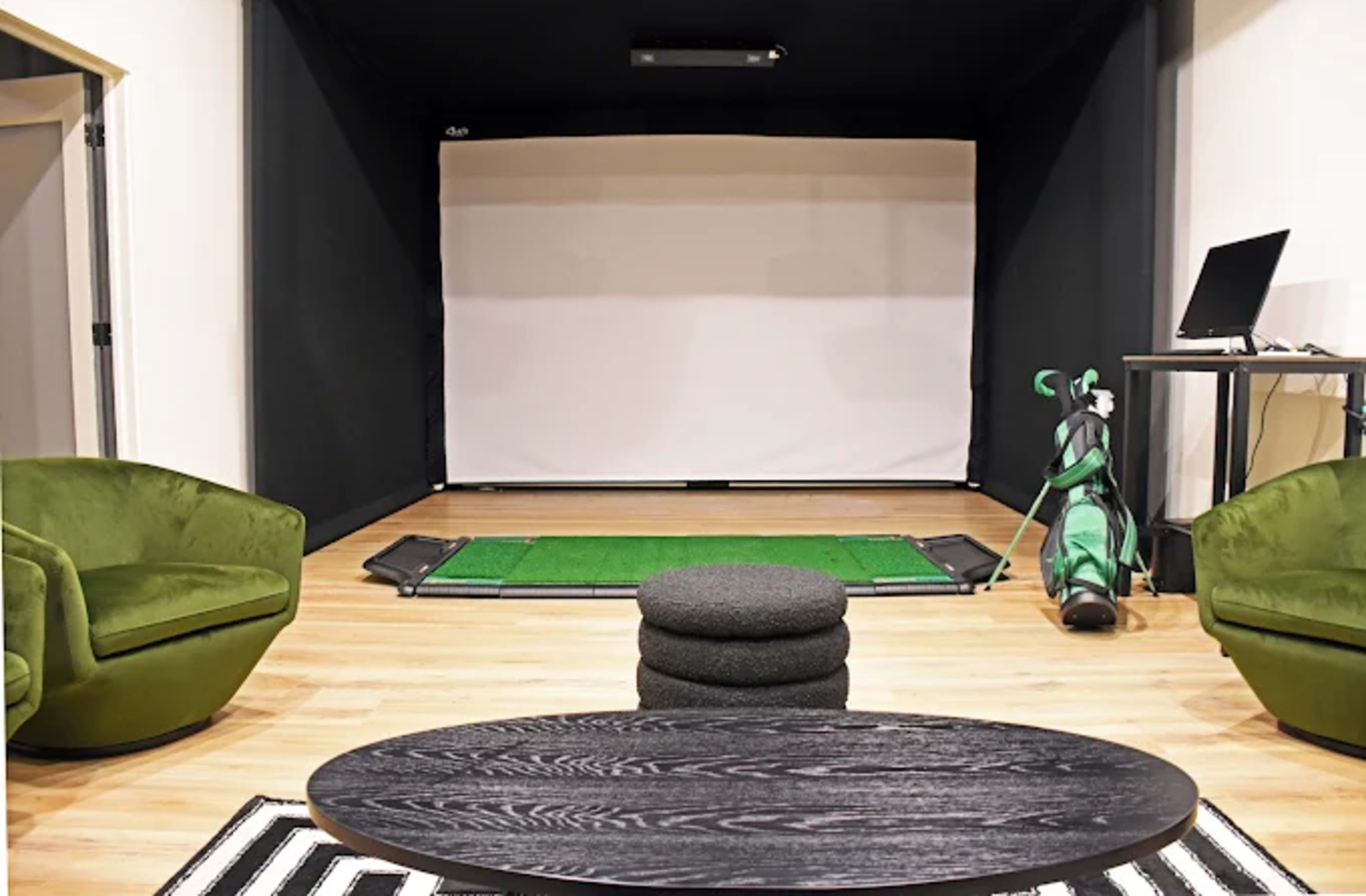 The image shows an indoor golf simulator setup featuring a large screen, a putting green, two green lounge chairs, a black coffee table, and a golf bag with clubs.