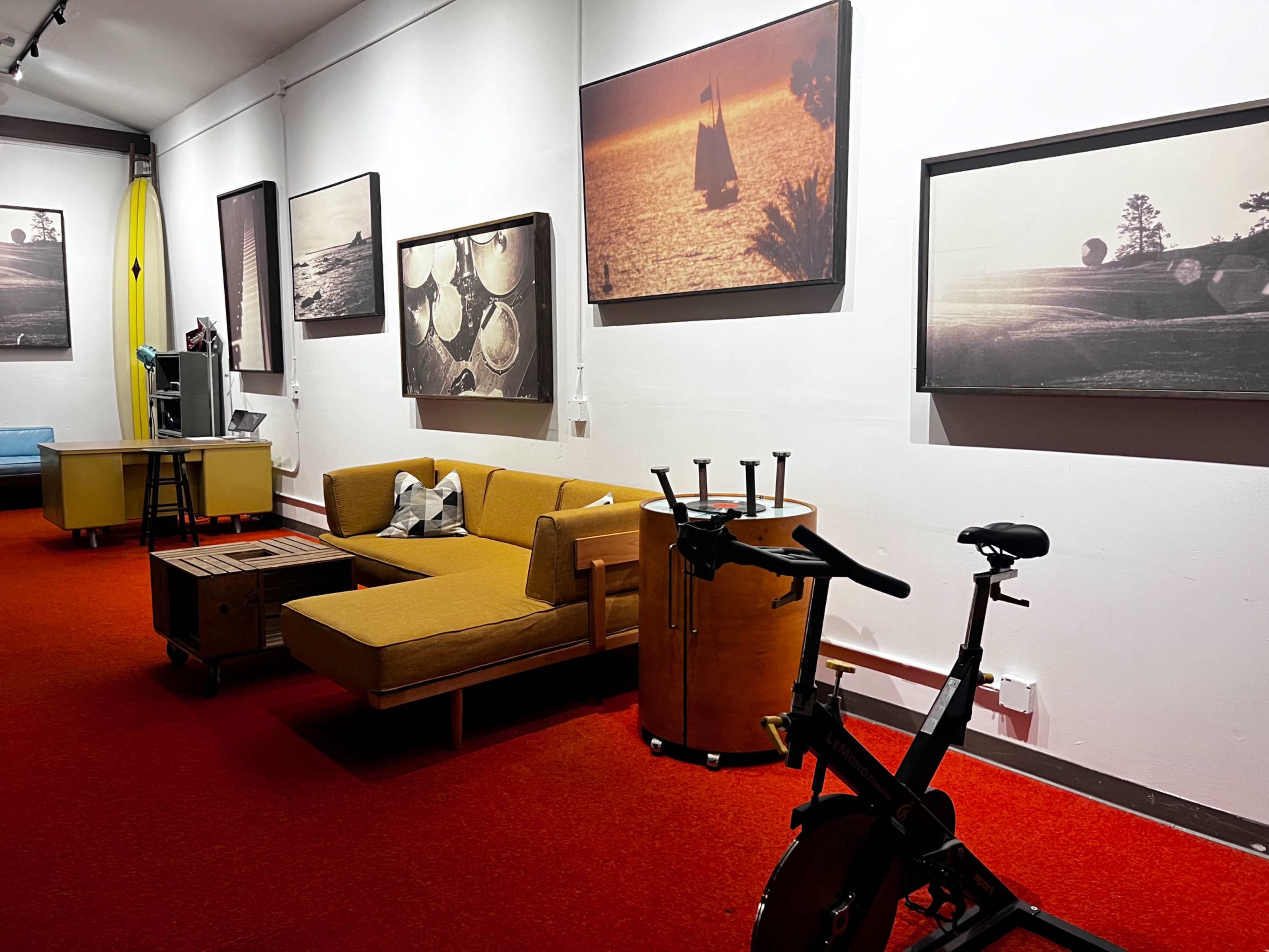 The image shows a room featuring a yellow sofa, a wooden table, and a stationary bike, with framed photographs hanging on the walls and red carpeting.