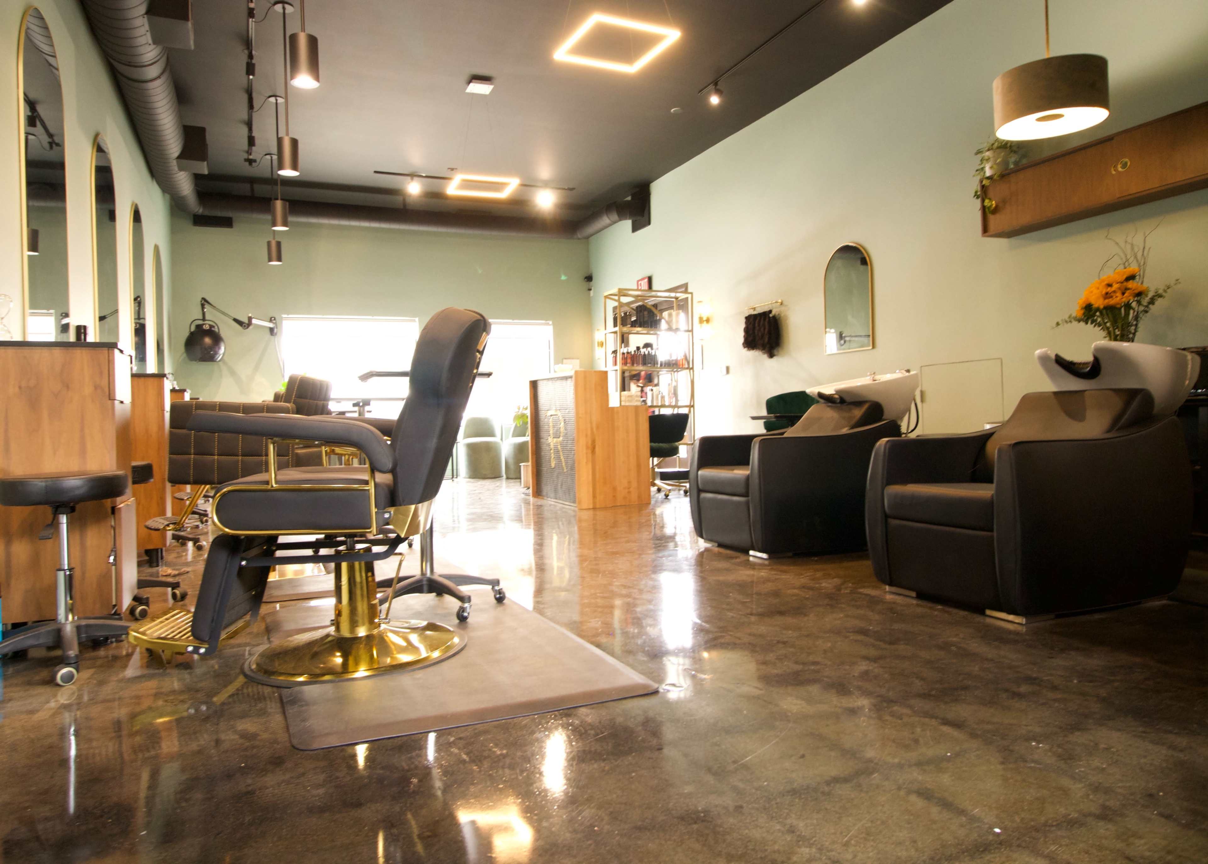 The interior of a hair salon features modern styling chairs, shampoo stations, and wooden furniture, with polished concrete flooring.