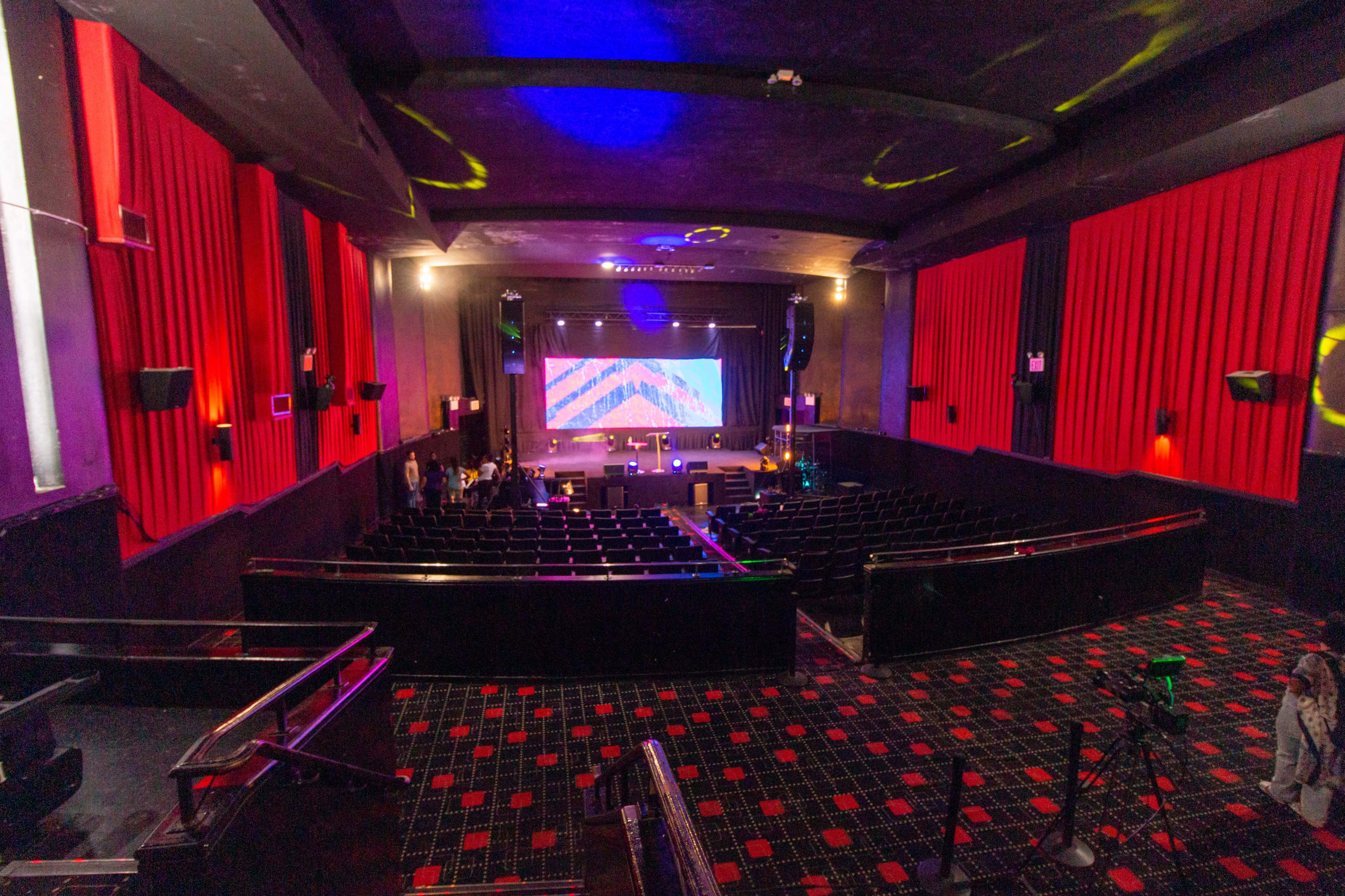 The interior of a theater with red curtains, a large screen at the front, and rows of empty black seats.