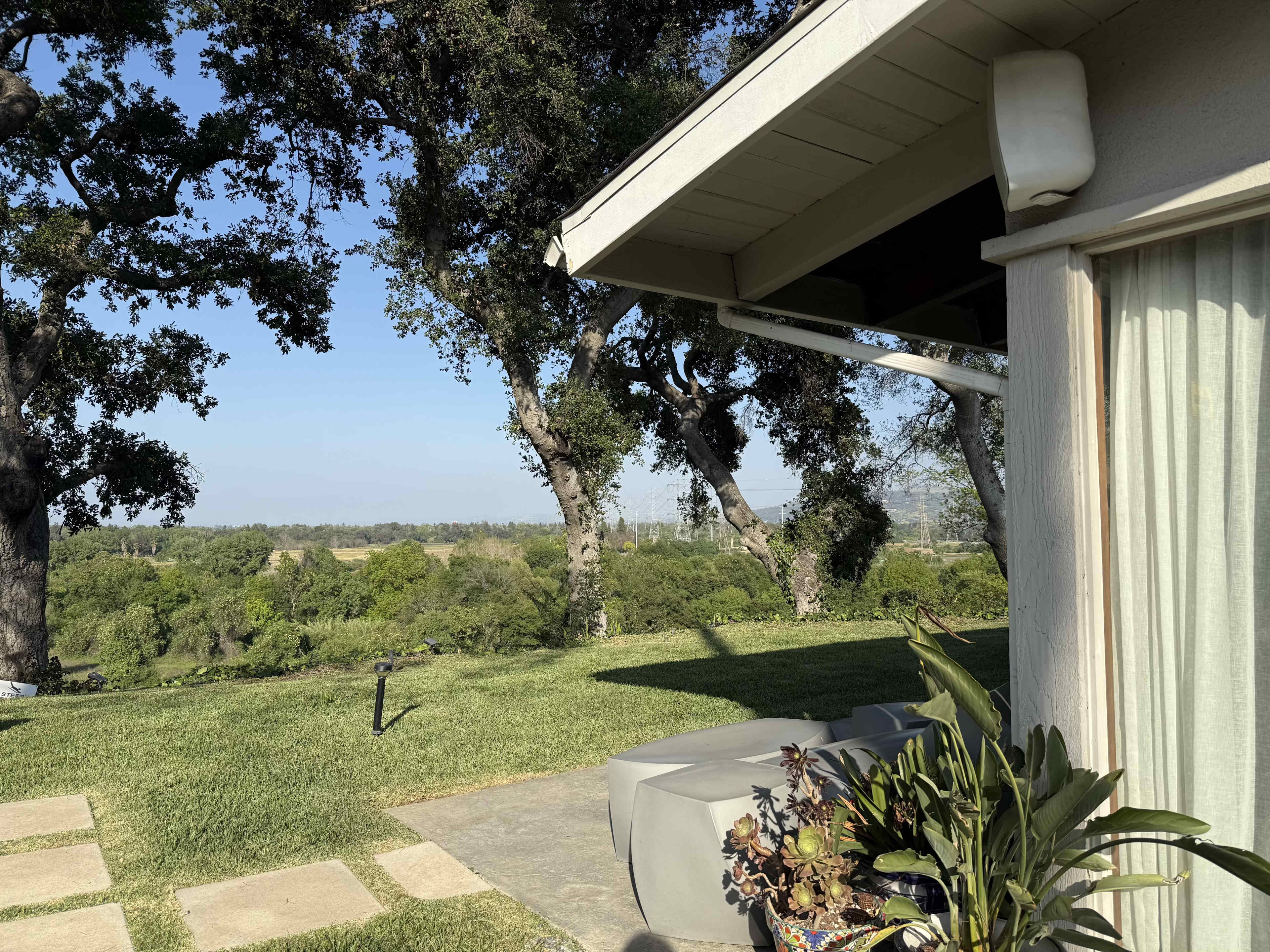 The image shows a landscaped yard with a stone pathway, green grass, and trees, alongside the exterior of a house featuring a large window and planter.