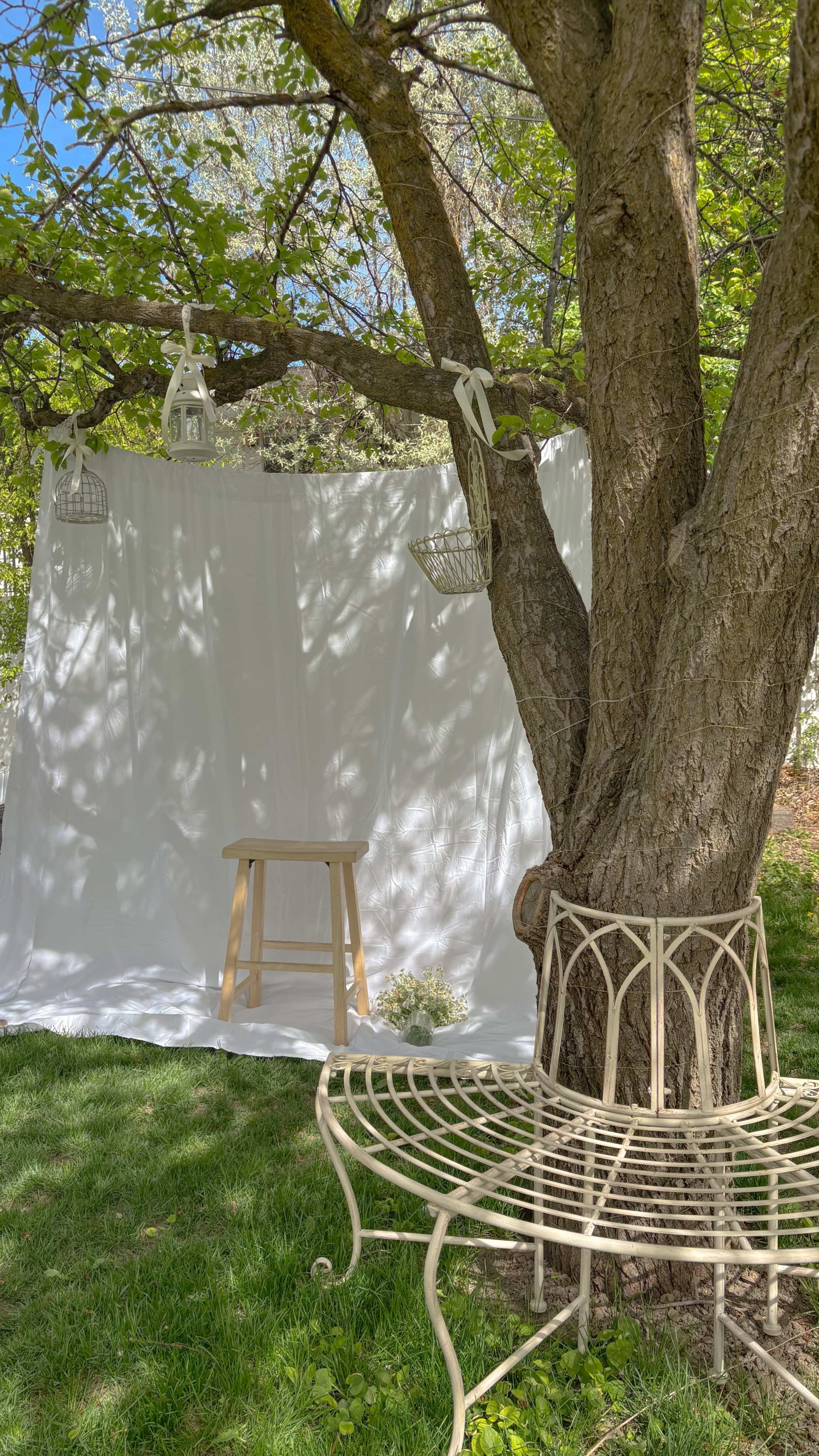 A white backdrop is draped between a tree and a stool, with hanging decorative elements above, set in a grassy area.