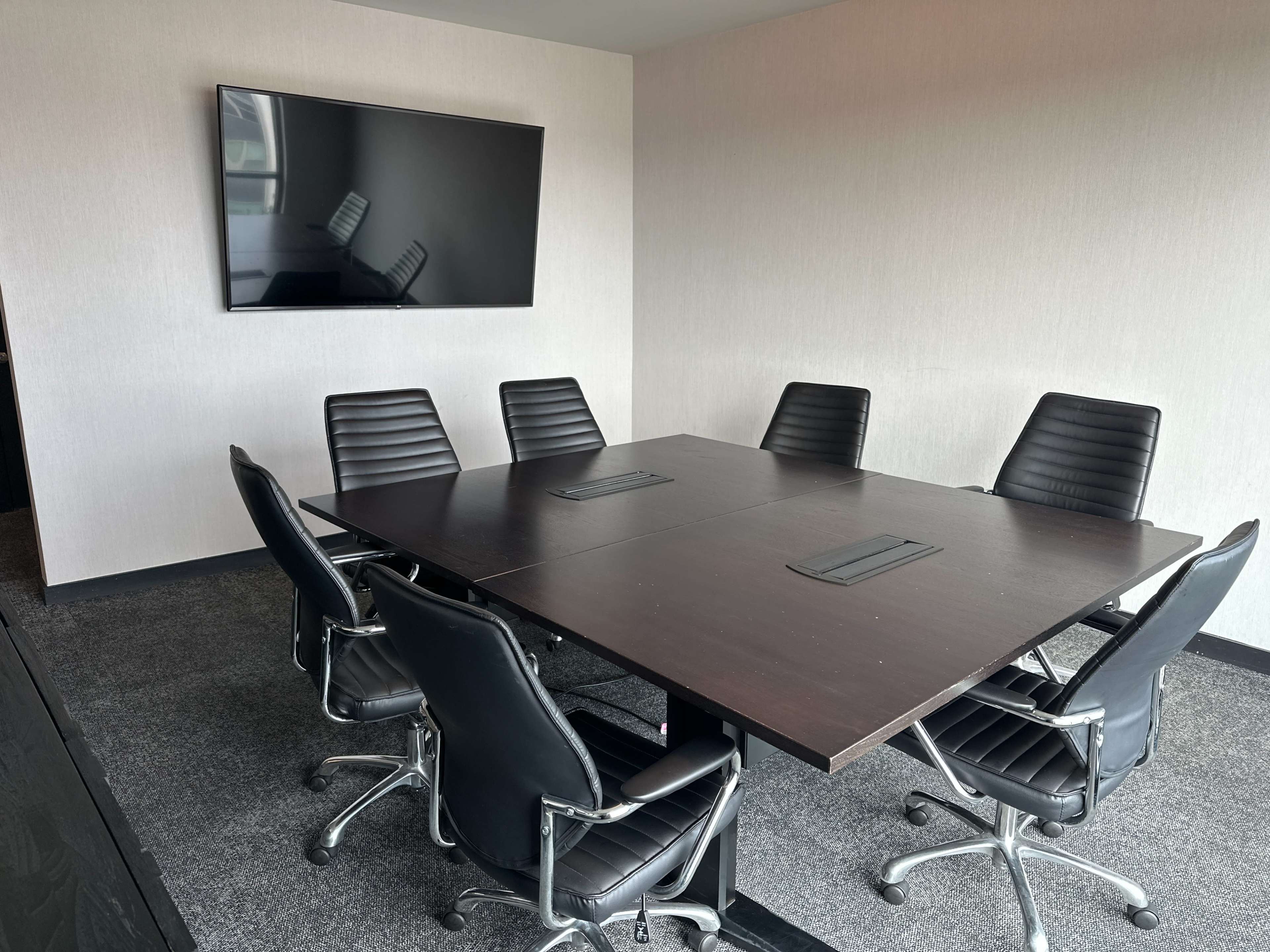 A modern conference room features a large wooden table surrounded by six black ergonomic chairs, with a wall-mounted television in the background.