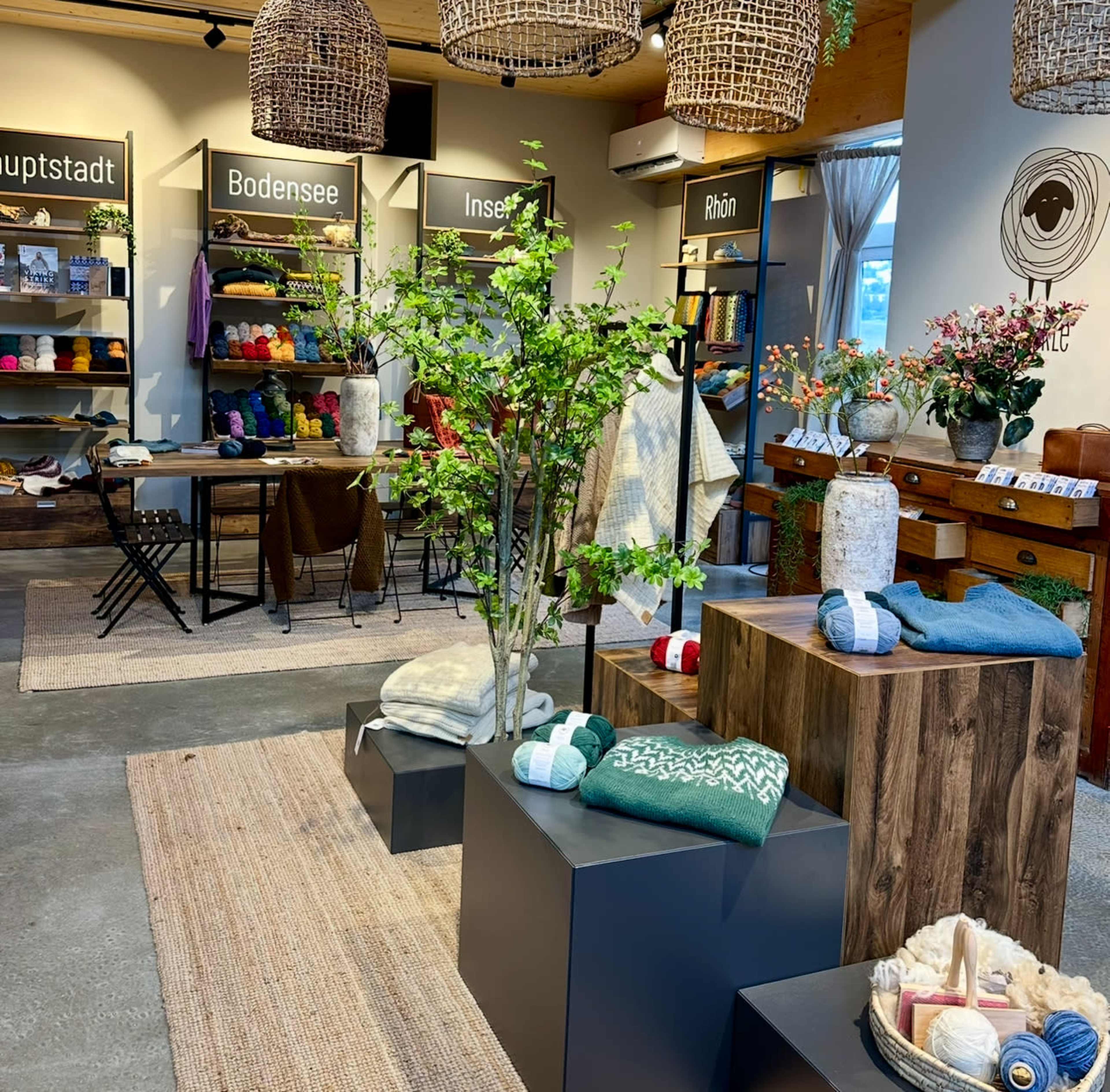 The image shows a cozy retail space featuring wooden displays with folded garments, greenery, and shelves filled with yarn, while tables for crafting are arranged in the background.