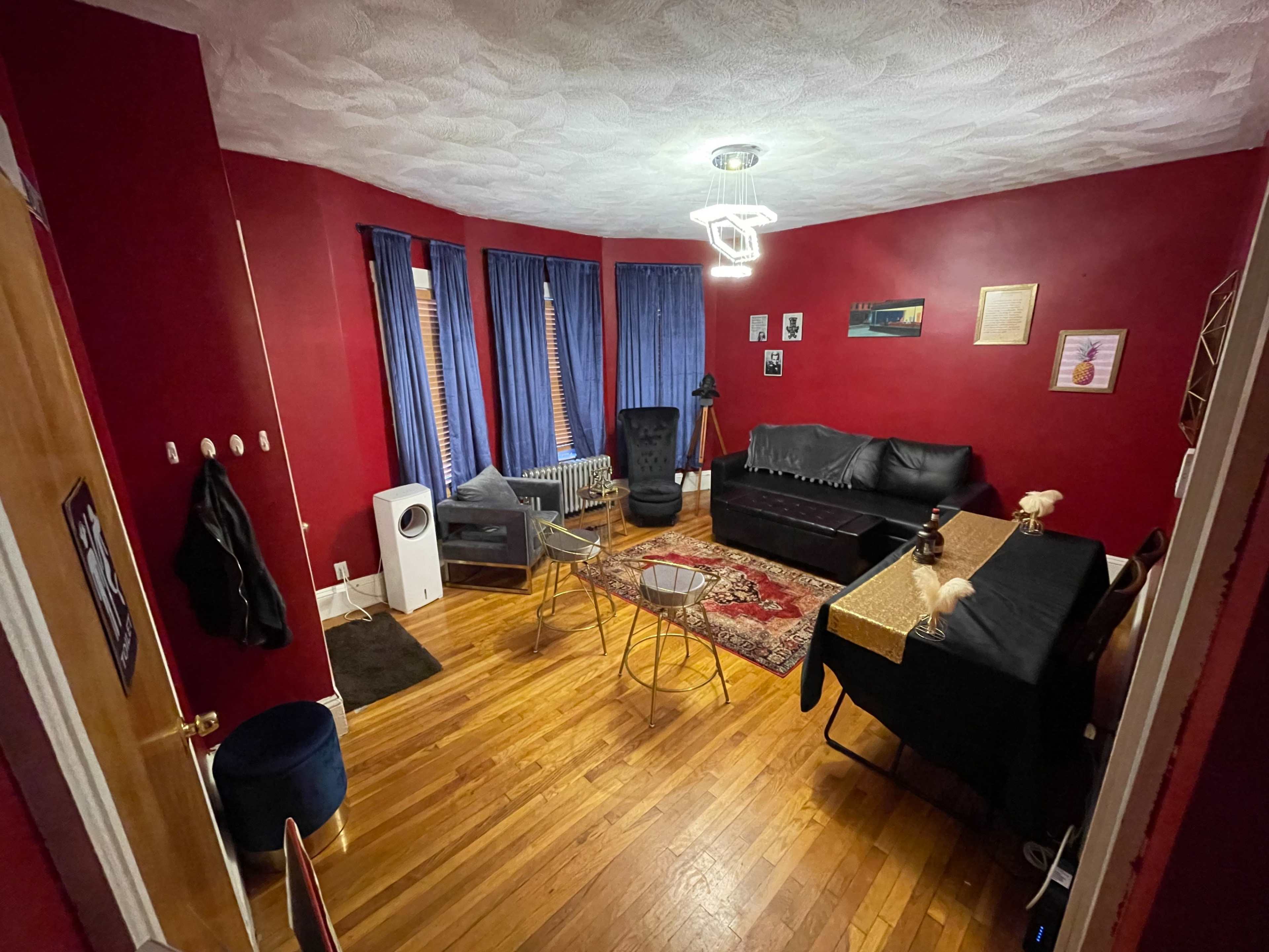 The room features red walls, wooden flooring, a black sofa, and a mix of seating along with a dining table set up.