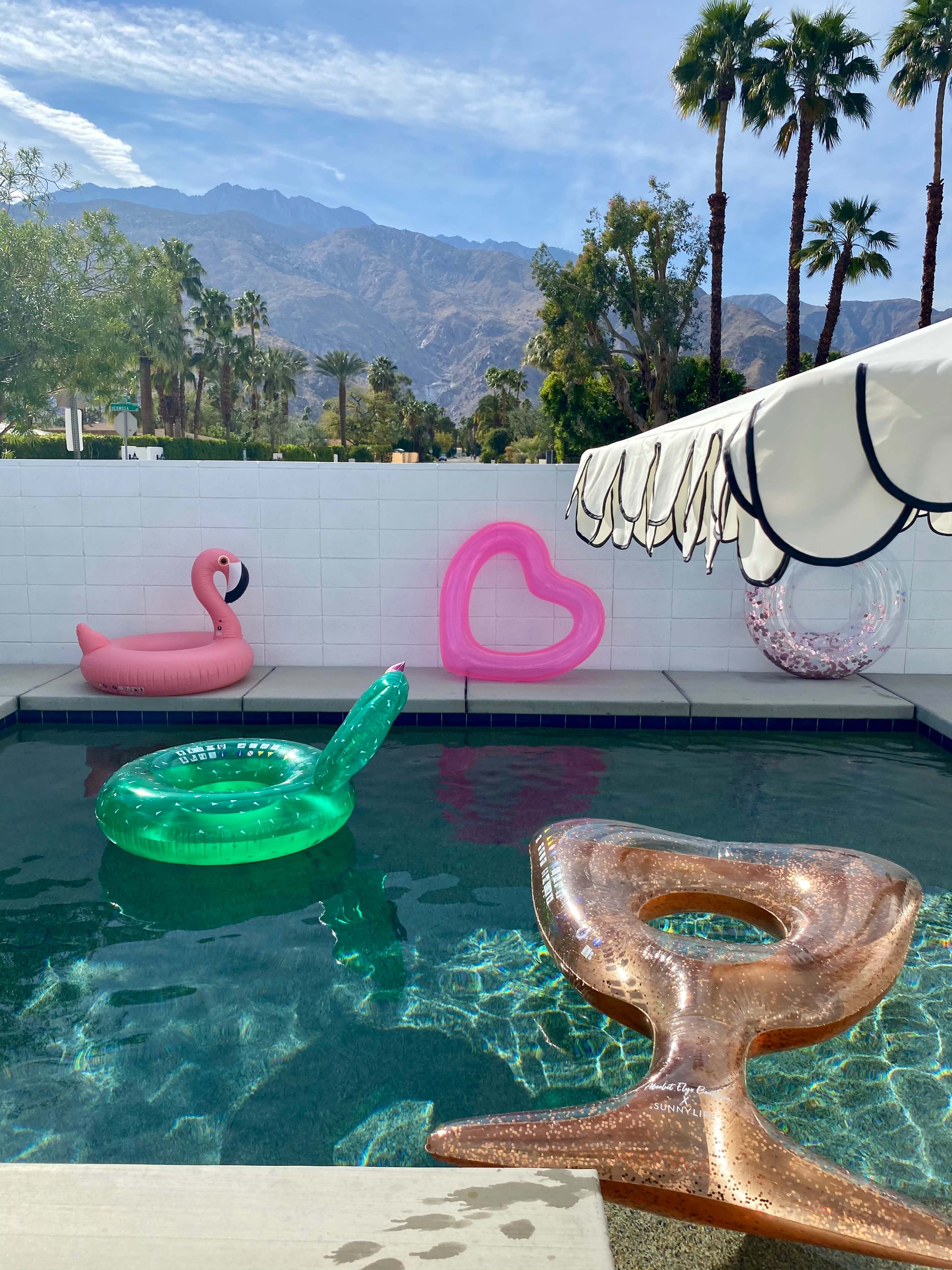 A swimming pool surrounded by inflatable toys, including a pink flamingo, a heart-shaped float, and a drink-shaped float, with palm trees and mountains in the background.