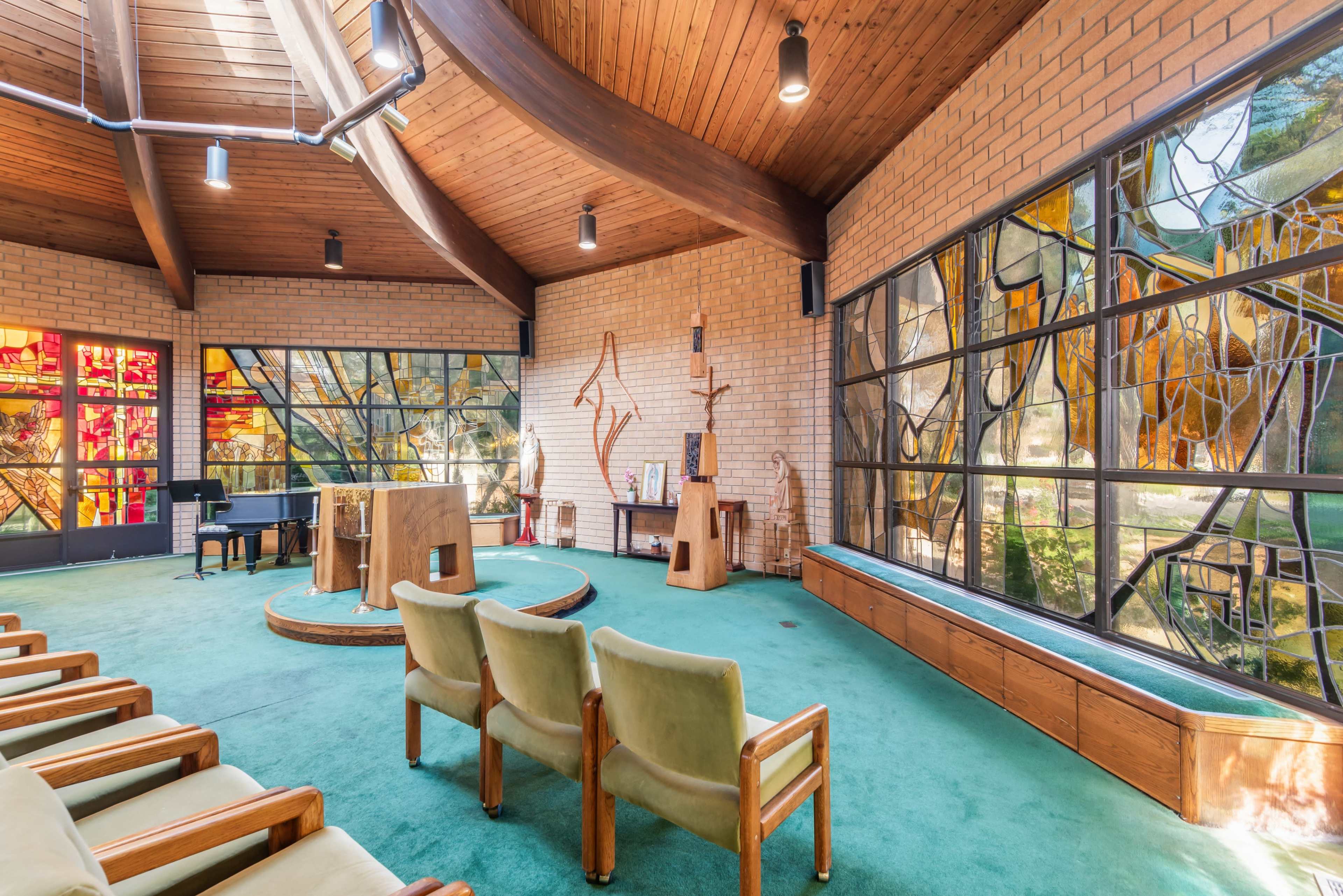 The image shows a spacious interior of a chapel with wooden architecture, colorful stained glass windows, and seating arranged in a semicircle around a central altar area.