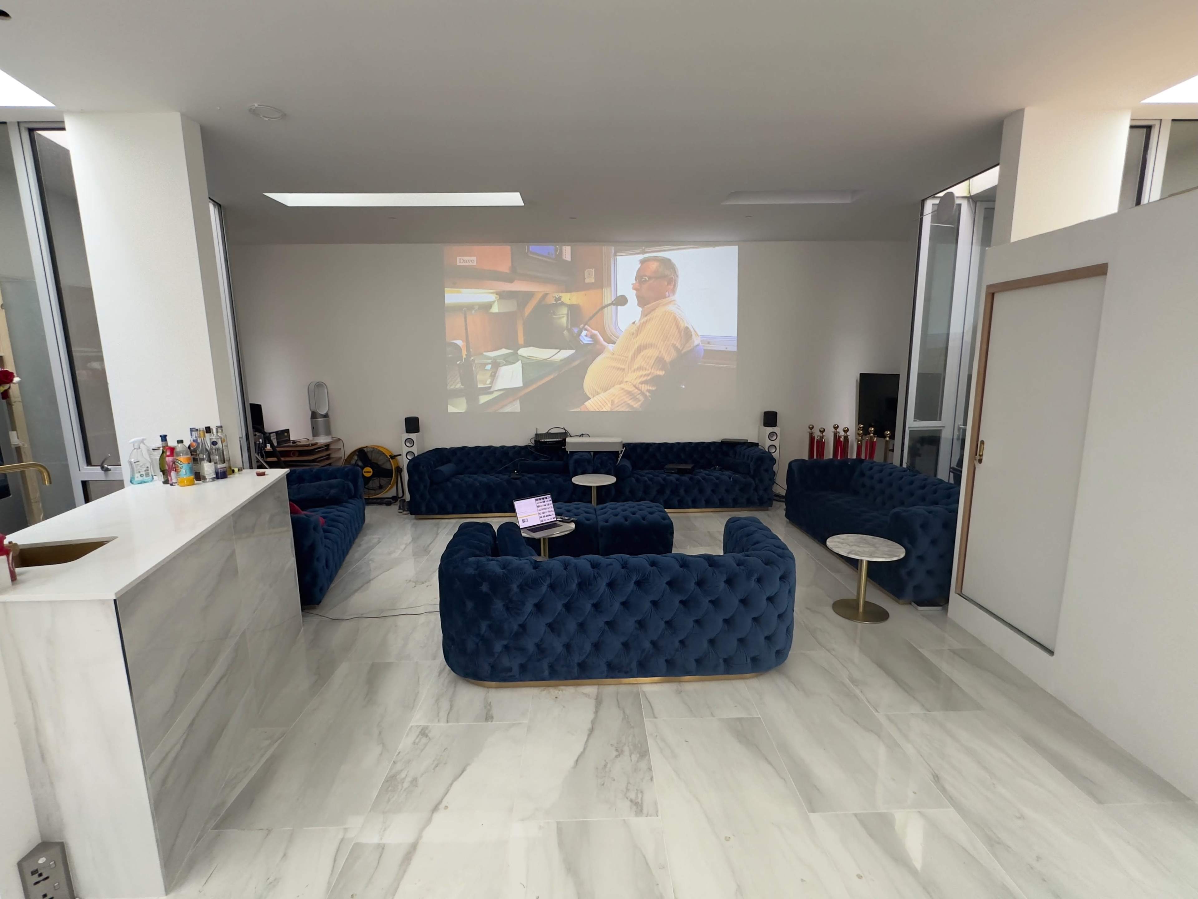 A modern lounge area features blue tufted sofas, a small round table, and a projector screen displaying a seated person.
