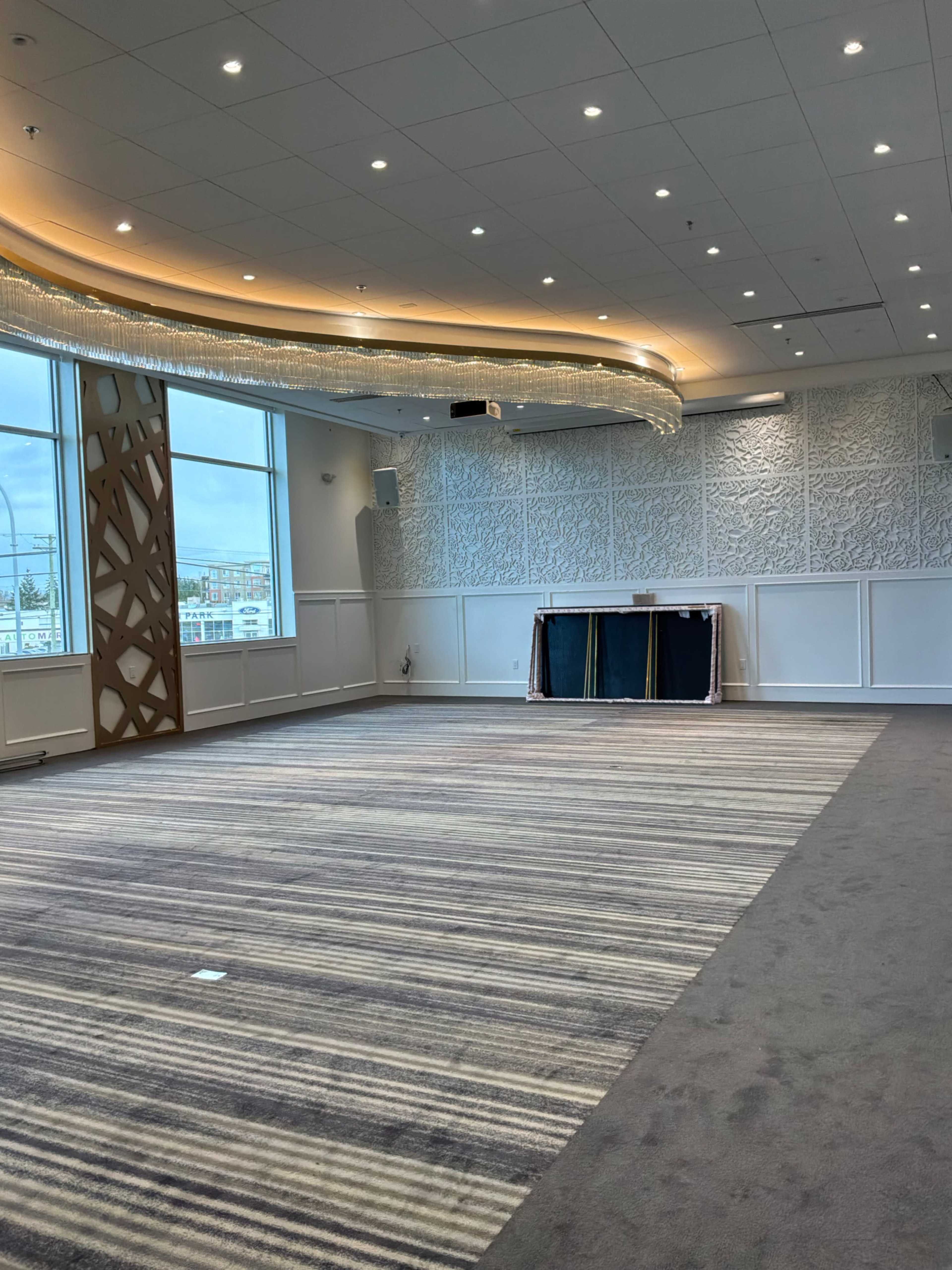 The image shows a spacious room with large windows, a patterned carpet, and a decorative light feature overhead.