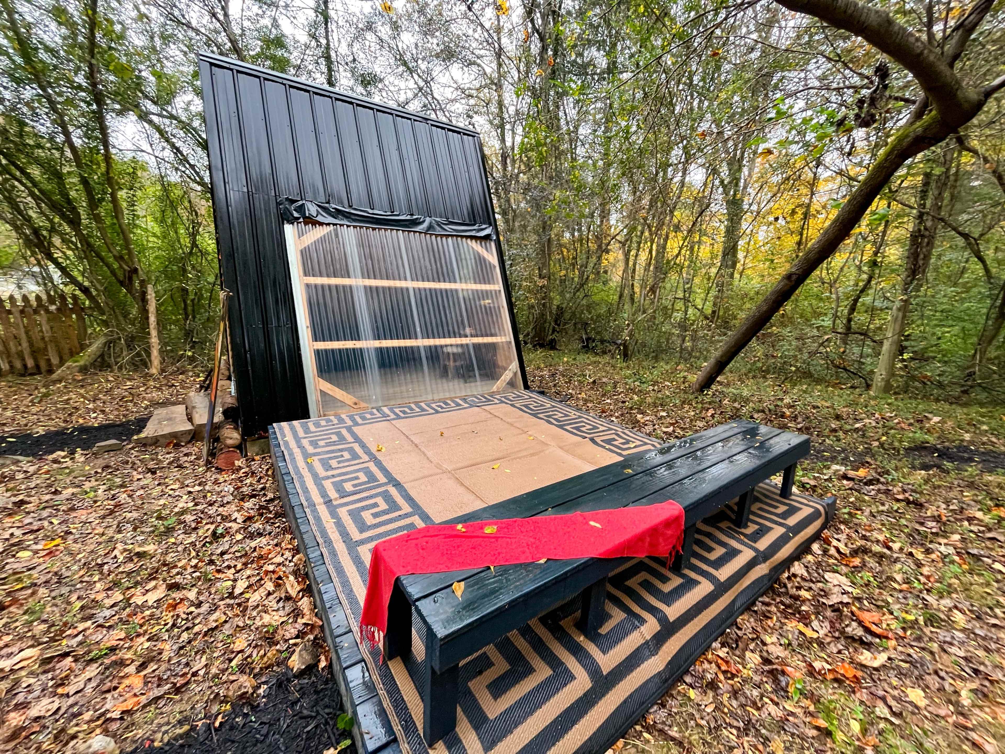 A black wooden structure with a transparent plastic covering, set on a patterned mat in a wooded area with fallen leaves.