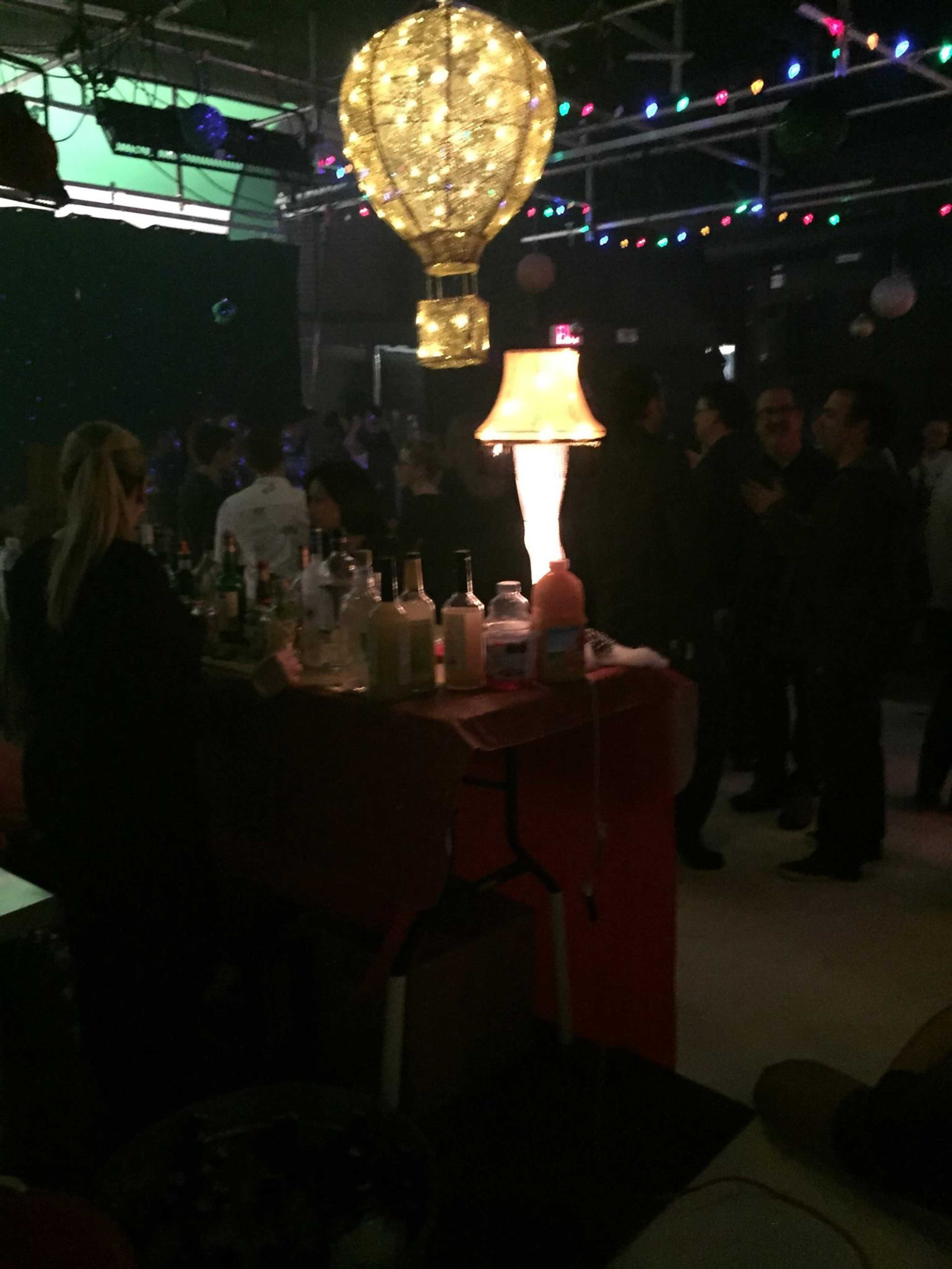 A bar area illuminated by a decorative lamp and surrounded by patrons in a dimly lit venue filled with colorful lights.