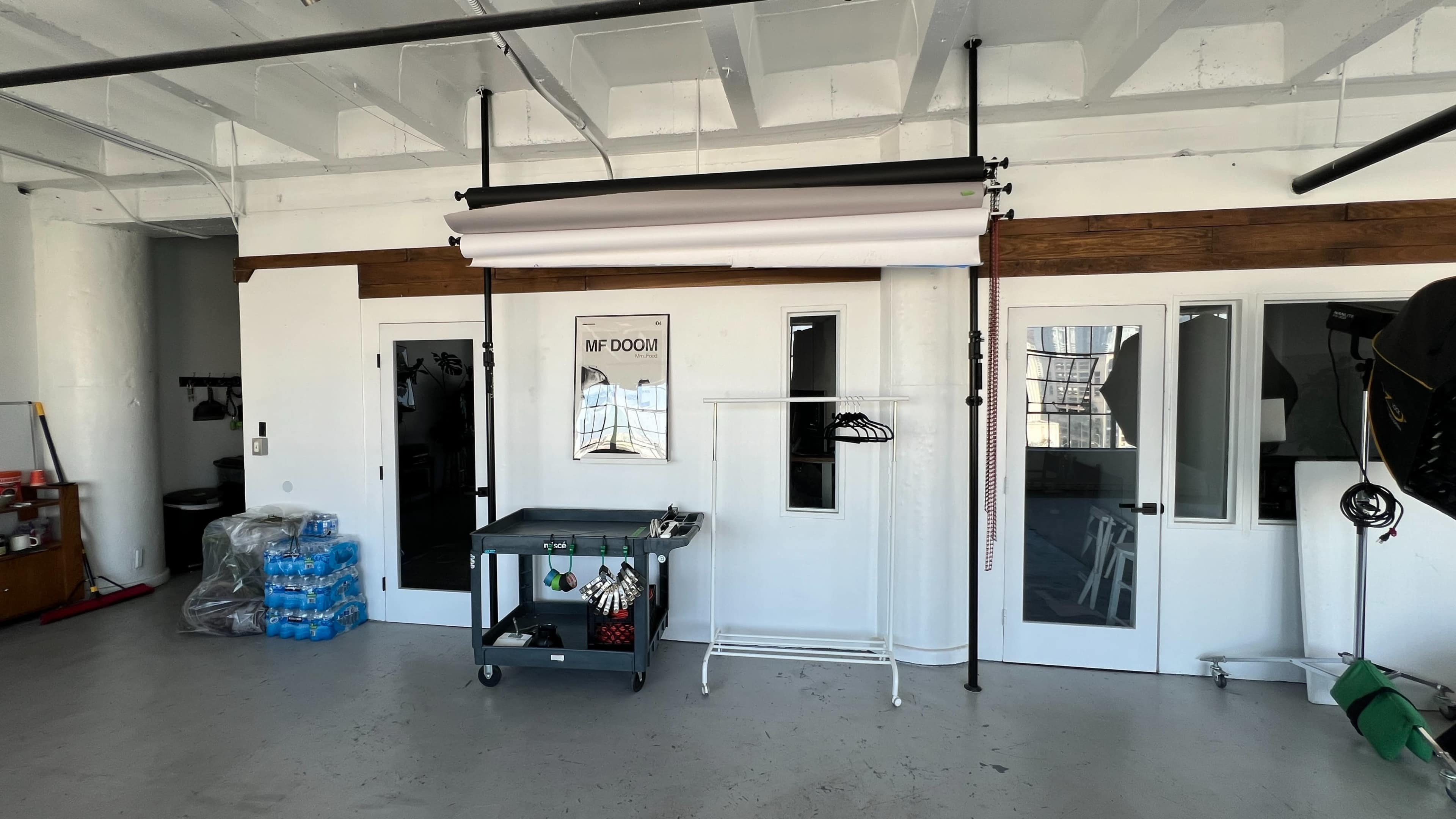 The image shows a spacious room with a rolling cart, a clothing rack, and large windows leading to a bright, open entrance area.