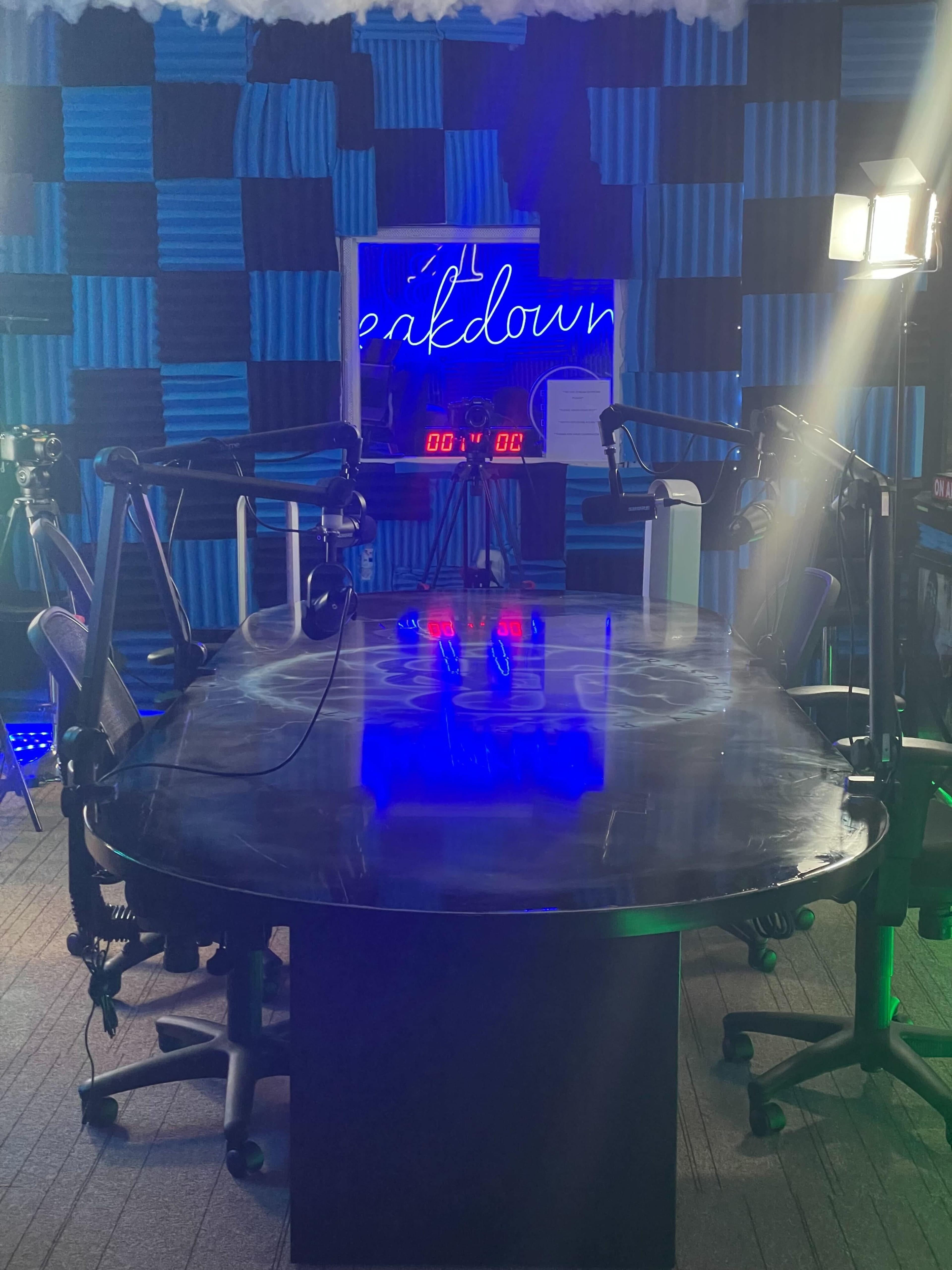 A recording studio features a large, glossy table surrounded by microphones, cameras, and colorful acoustic wall panels.