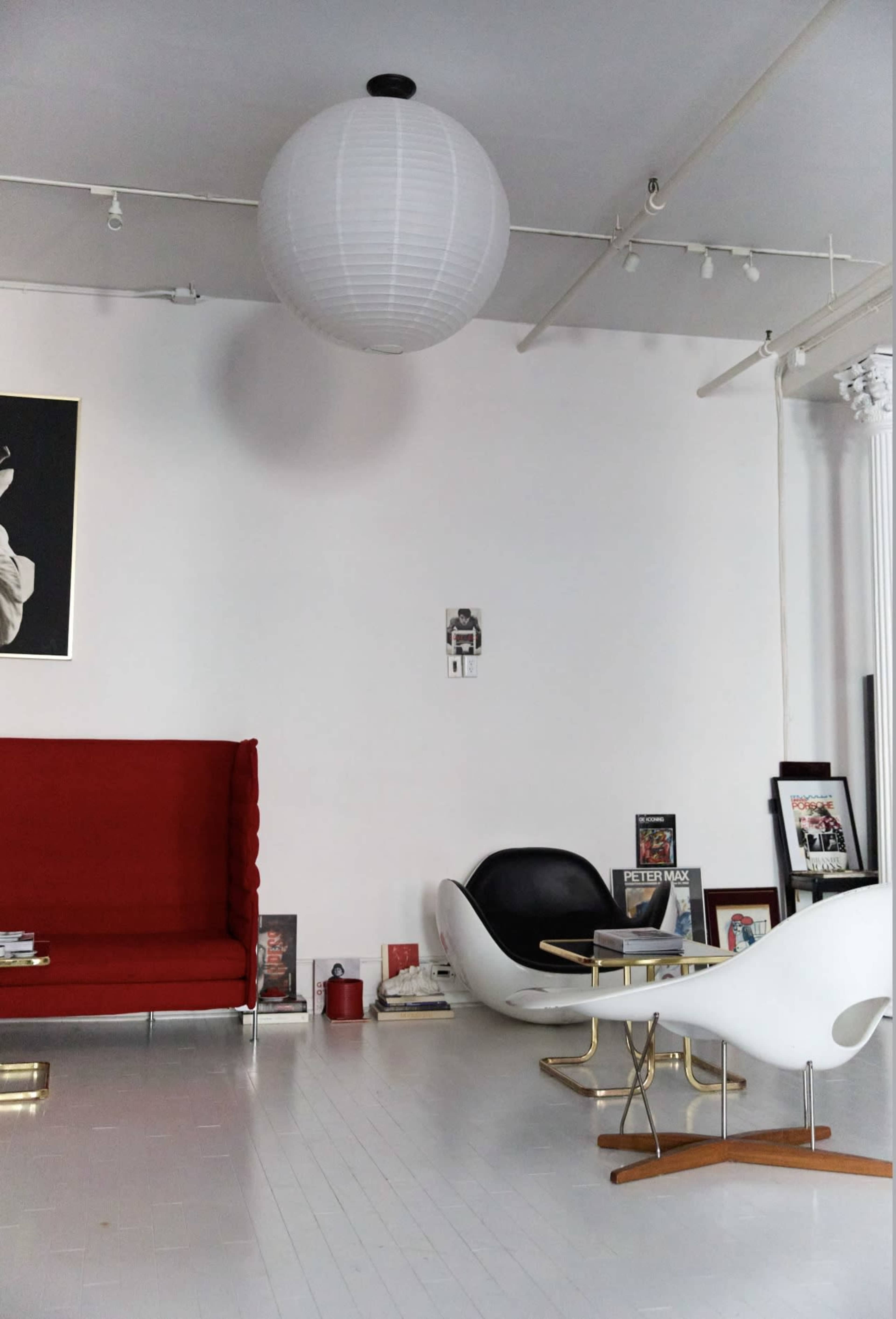The image shows a modern interior featuring a red couch, a black and white chair, a paper lantern ceiling light, and framed pictures against a white wall.