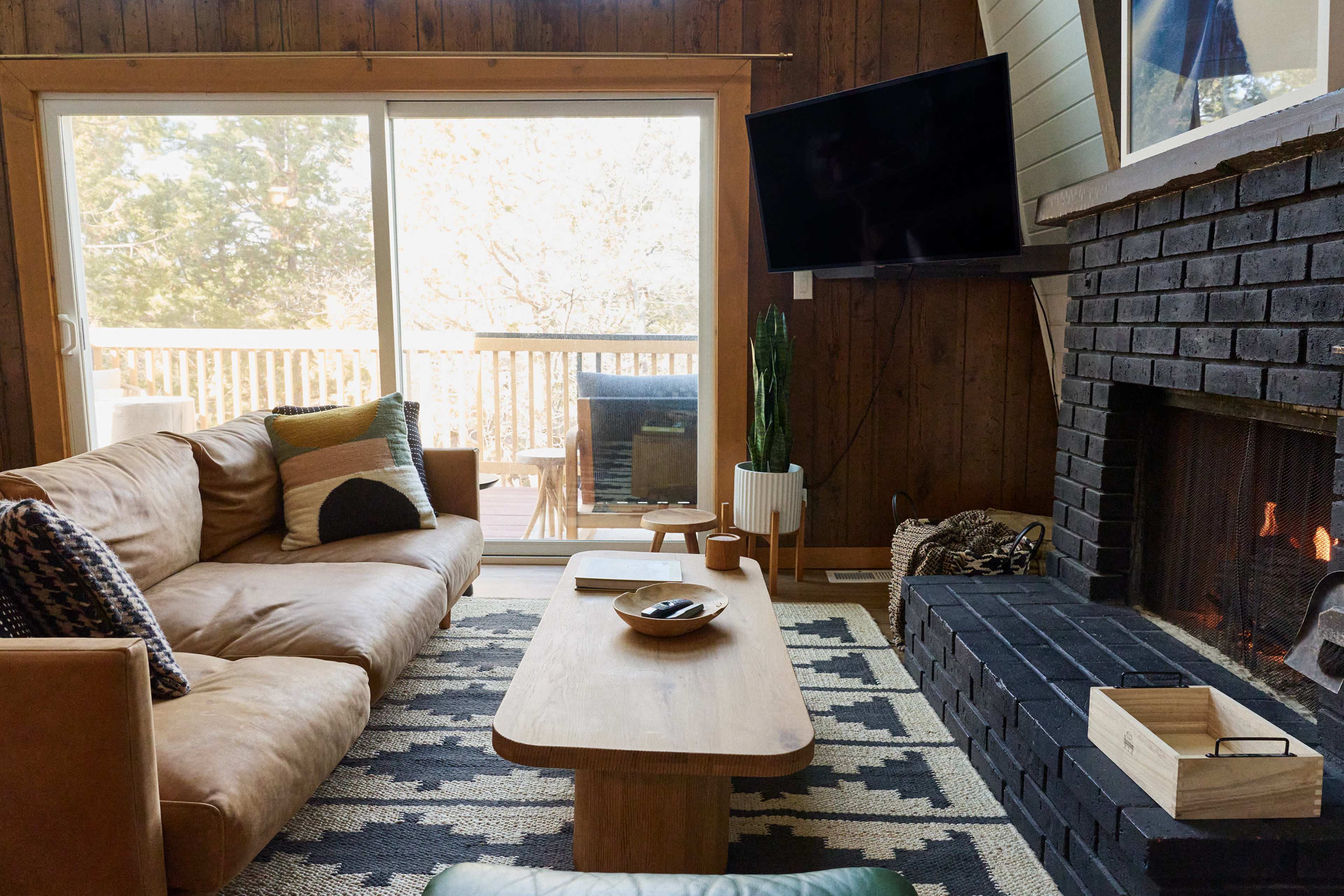 The image shows a cozy living room with a brown leather sofa, a wooden coffee table, a black brick fireplace, and a large sliding glass door leading to an outdoor deck.