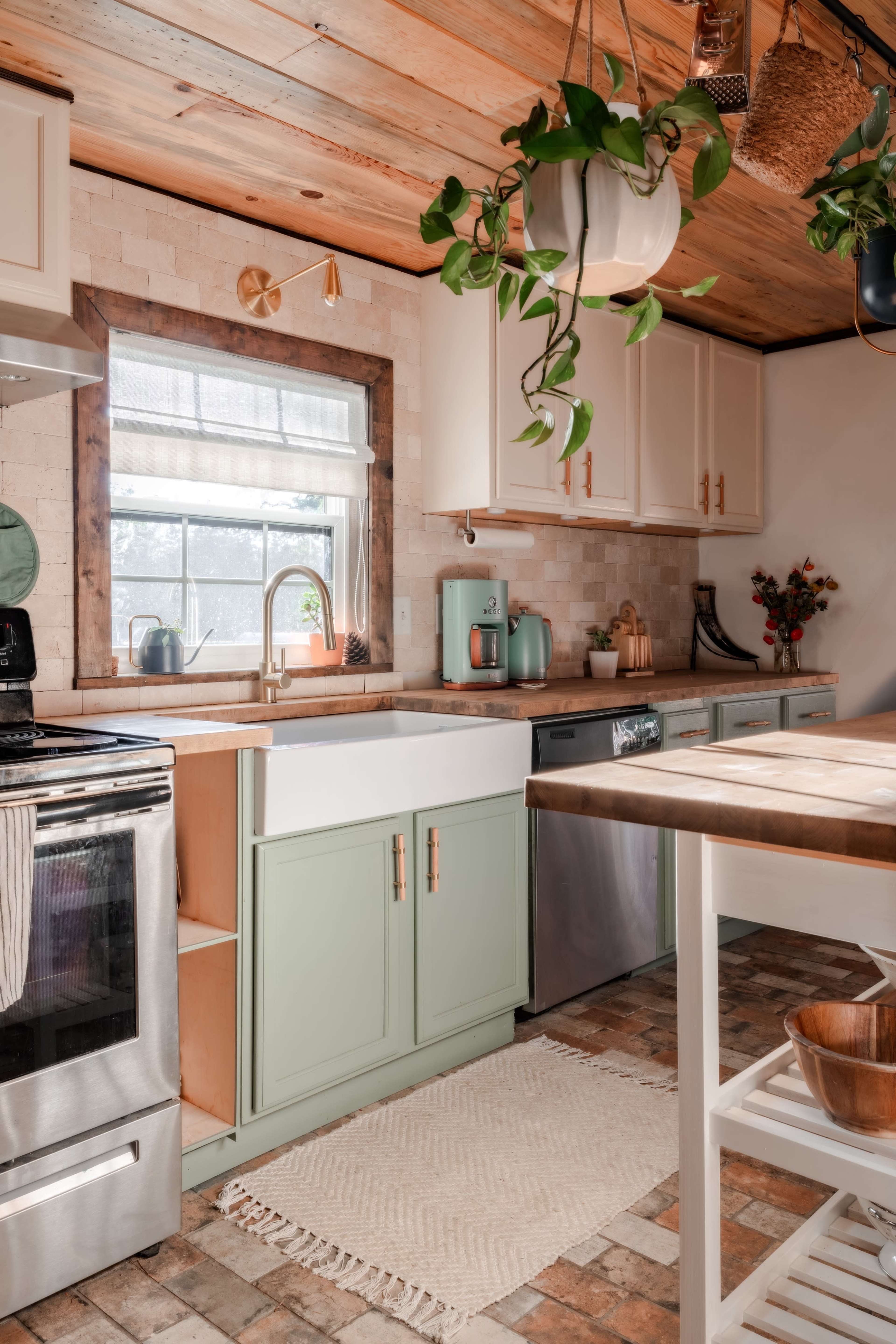 A modern kitchen with a large farmhouse sink, stainless steel appliances, and green cabinetry, complemented by hanging plants and natural wood accents.