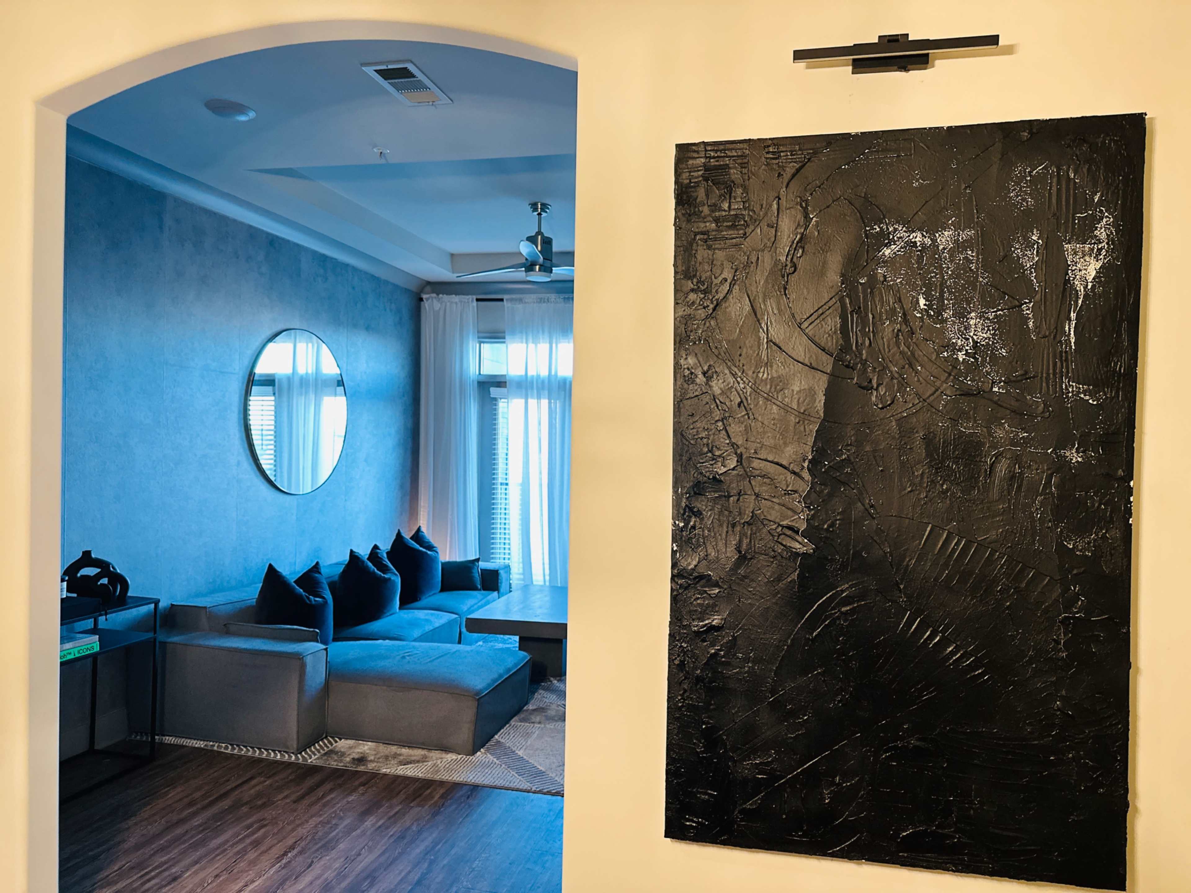 A modern living room with a circular mirror and a gray sectional sofa is visible through an arched doorway, next to a large textured black artwork mounted on the wall.