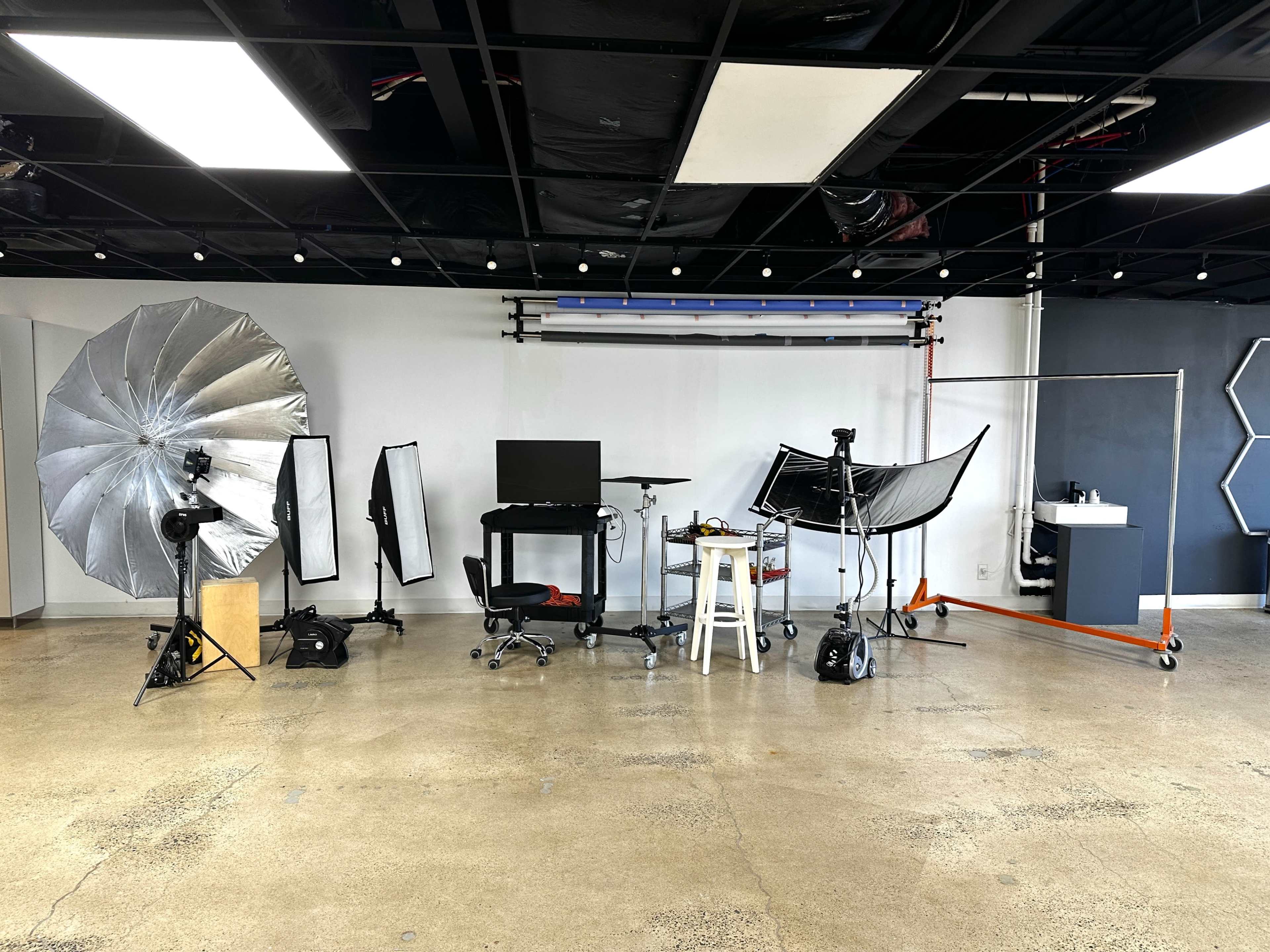 The image shows a photography studio setup with various lighting equipment, backgrounds, and a computer workstation arranged on a concrete floor.