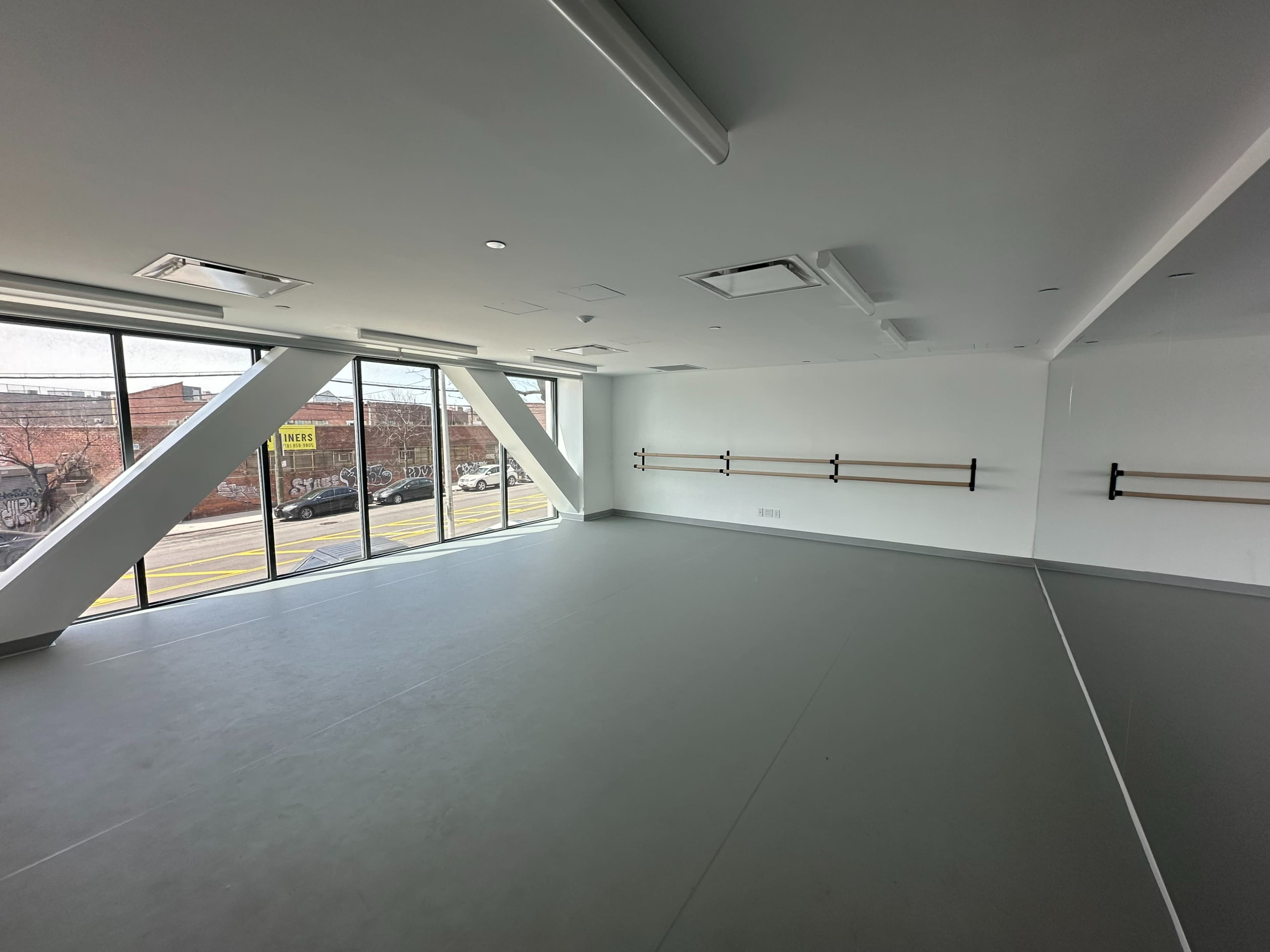 The image shows a spacious, empty studio with large windows and mirrored walls, featuring ballet bars along one side.