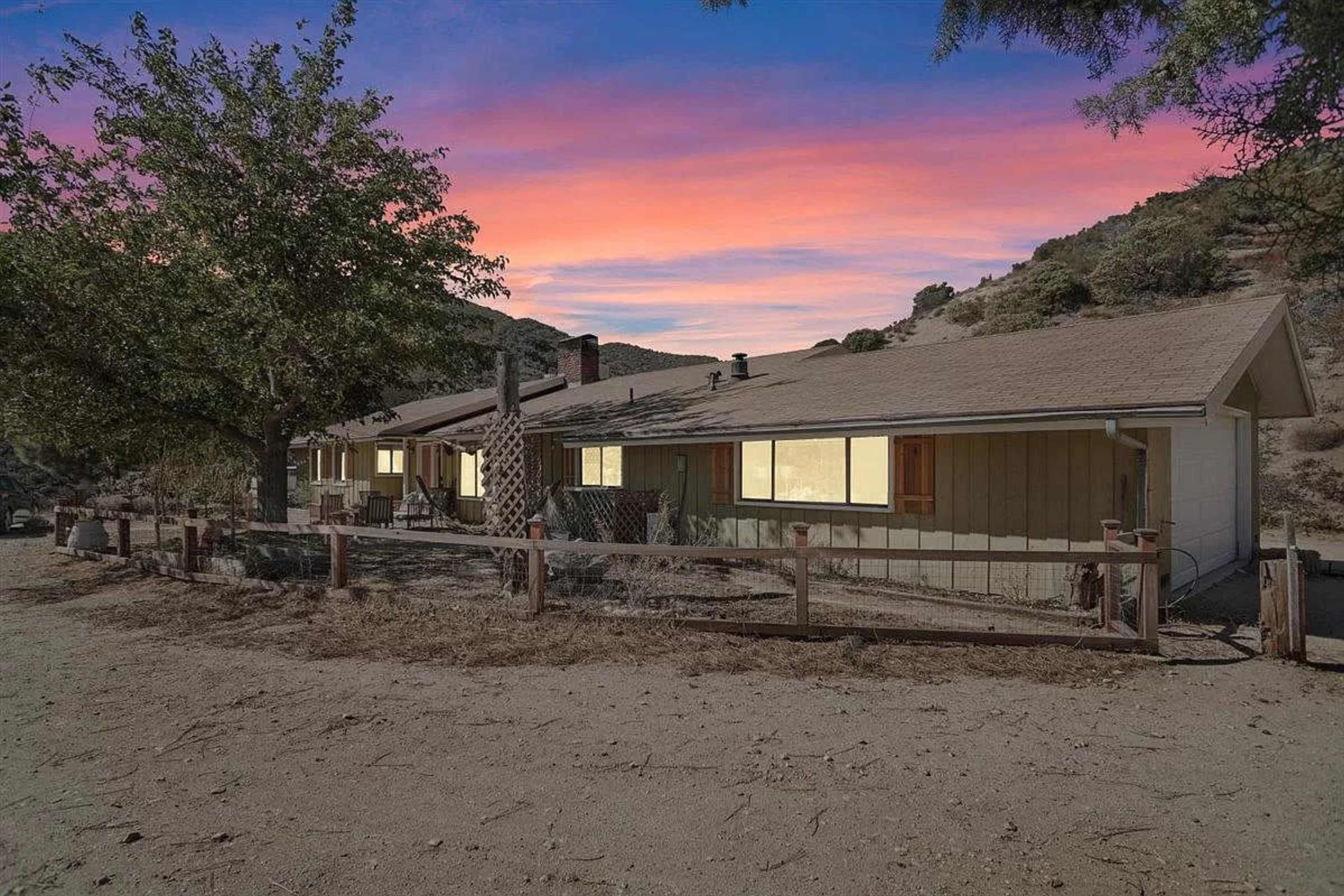 A single-story house with a fenced yard is situated in a mountainous area, surrounded by sparse vegetation under a colorful sunset sky.