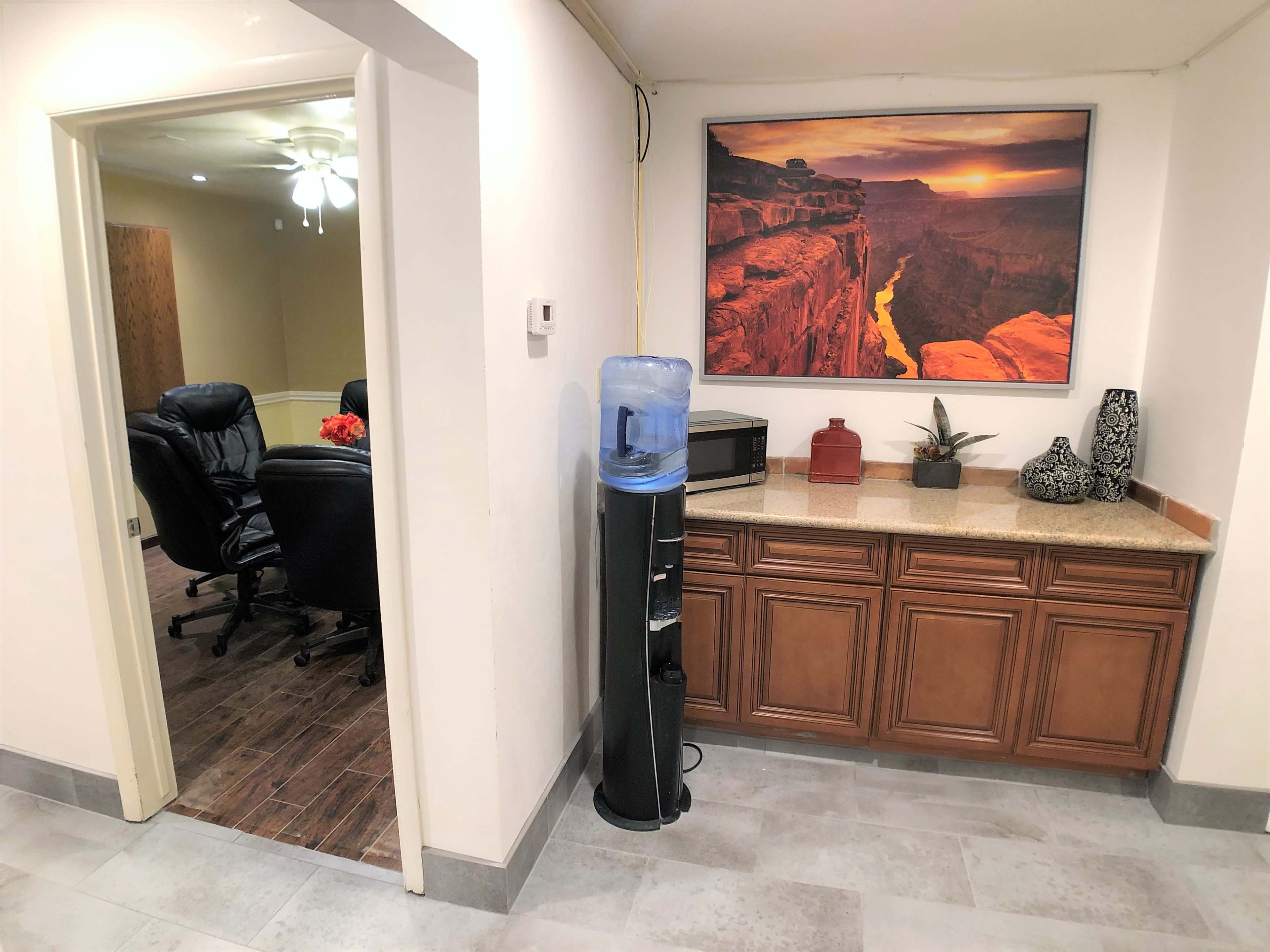 The image shows a small office space featuring a water cooler next to a countertop with decorative items, and a doorway leading to a room with black chairs and a ceiling fan.
