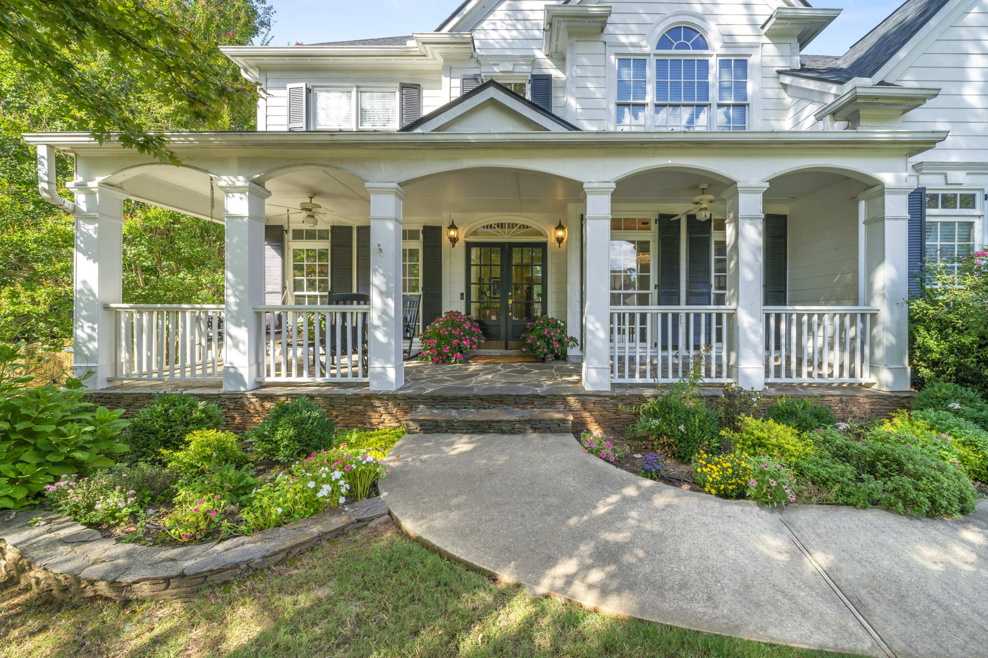 A two-story house with a large front porch, surrounded by colorful flower beds and a stone walkway leading to the entrance.